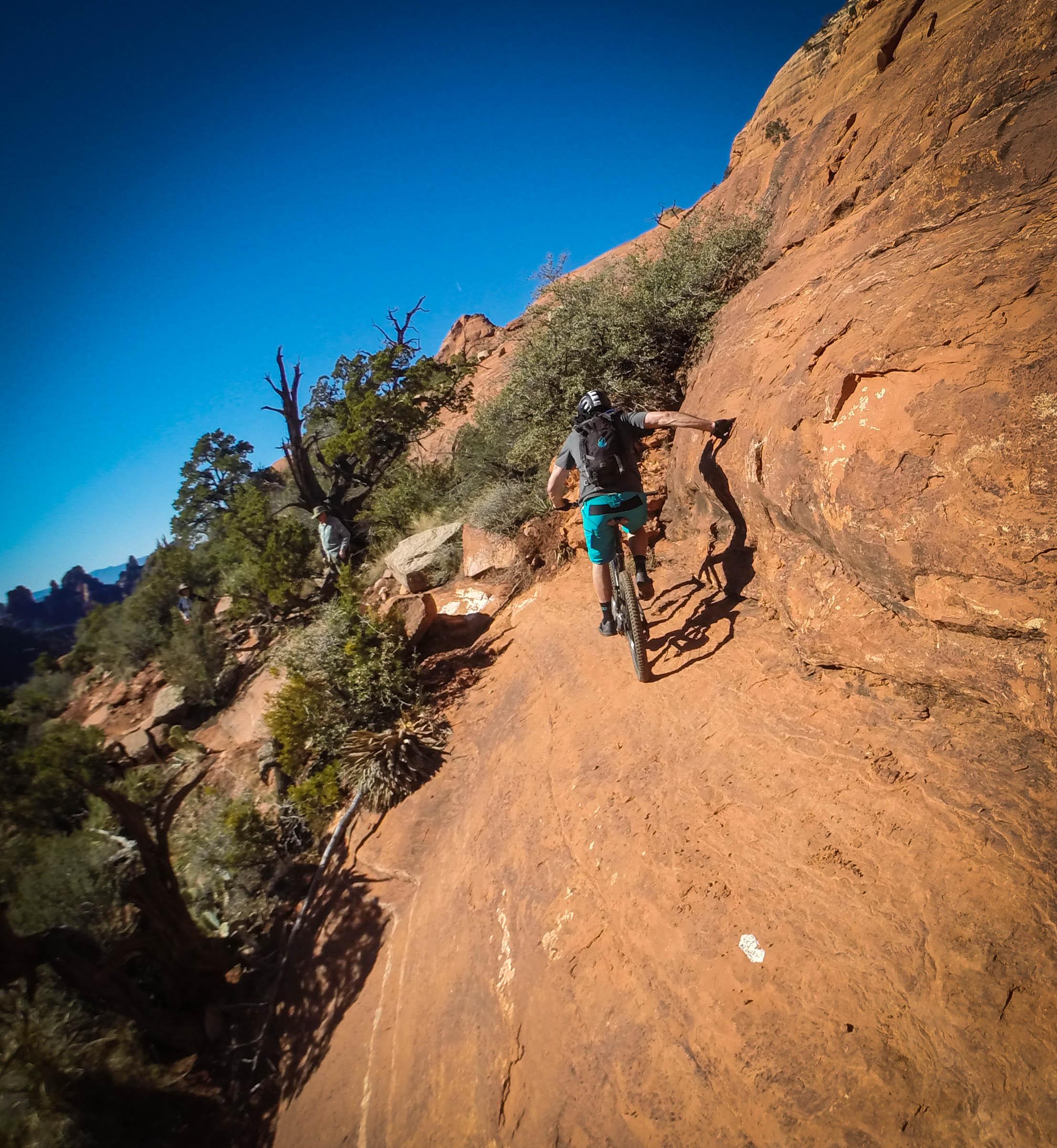 A person riding a mountain bike on a narrow, rocky trail surrounded by desert vegetation and a clear blue sky. The terrain features reddish-brown rocks and sparse trees, with distant rock formations visible in the background. Hangover mountain bike trail.