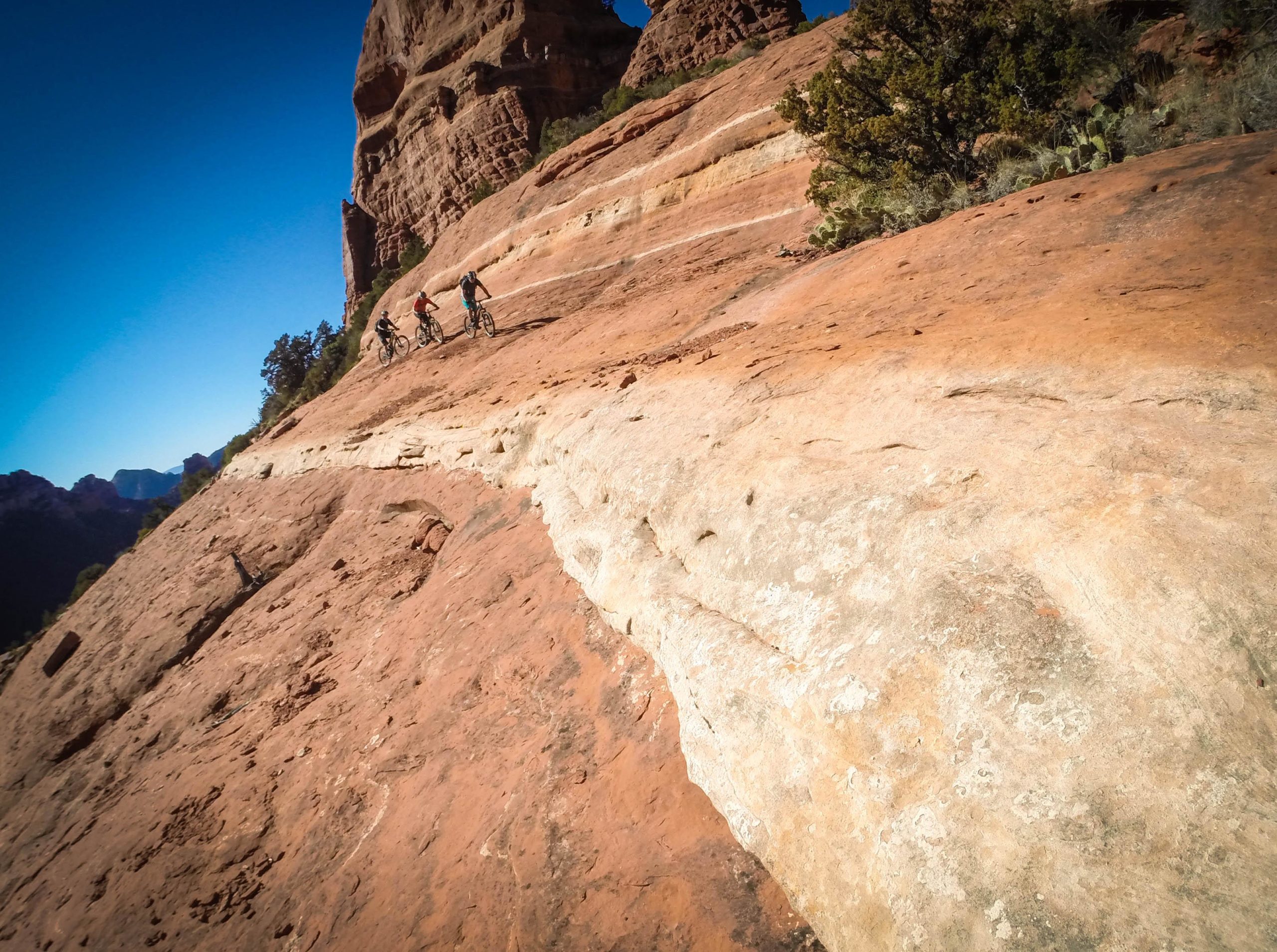 Three mountain bikers riding on a rocky, sloped terrain surrounded by red rock formations under a clear blue sky. Hangover mountain bike trail.