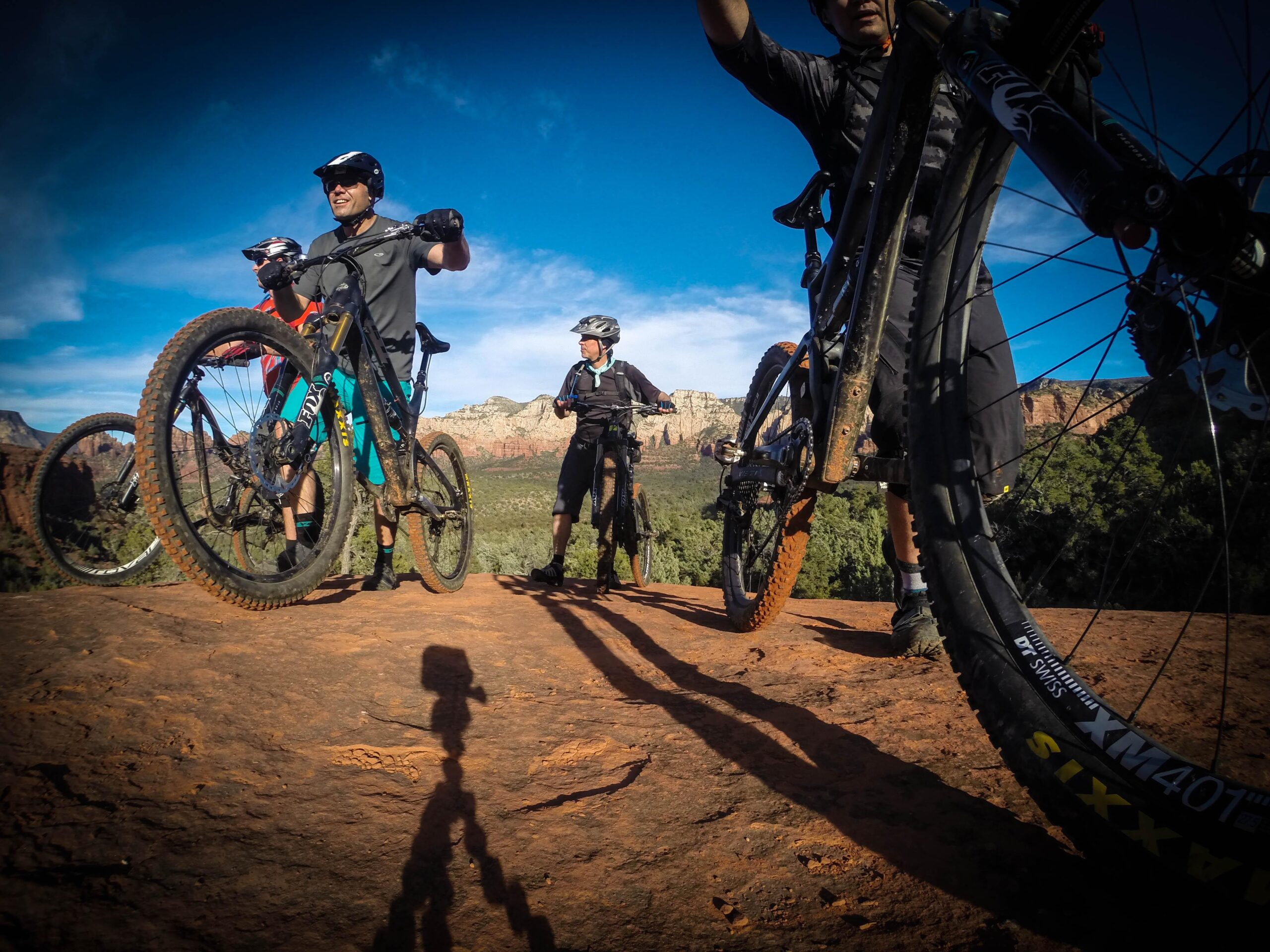 A group of four mountain bikers on a rocky outcrop, each holding their bikes and overlooking a scenic landscape. The sky is clear with blue hues, and red rock formations are visible in the background. The bikers are dressed in athletic gear, and the terrain is rugged and natural. Broken Arrow Trail / Chicken Point mountain bike trail.