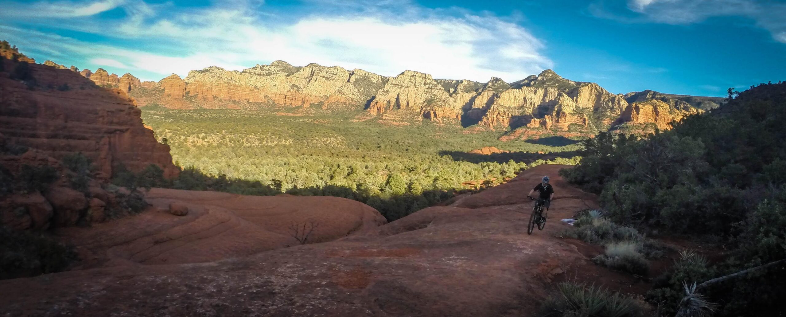 A mountain biker rides along a rocky trail with scenic red rock formations and green vegetation in the background under a clear blue sky. The landscape features rugged mountains and a valley, showcasing the natural beauty of the outdoors. Broken Arrow Trail / Chicken Point mountain bike trail.