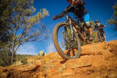 Two mountain bikers navigating rocky terrain, with one rider descending a steep section of red earth while the other follows closely behind. The scene is set against a bright blue sky and surrounded by sparse greenery and cacti. Chuck Wagon mountain bike trail.