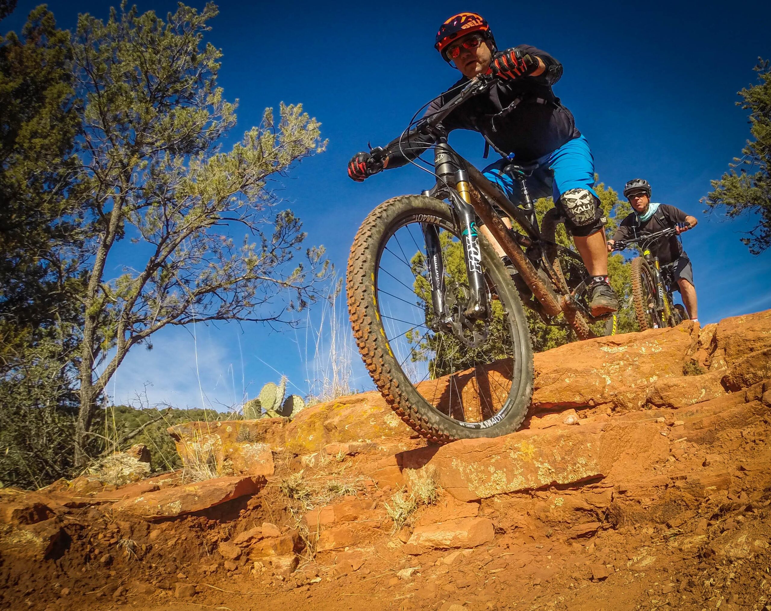Two mountain bikers navigating rocky terrain, with one rider descending a steep section of red earth while the other follows closely behind. The scene is set against a bright blue sky and surrounded by sparse greenery and cacti. Chuck Wagon mountain bike trail.
