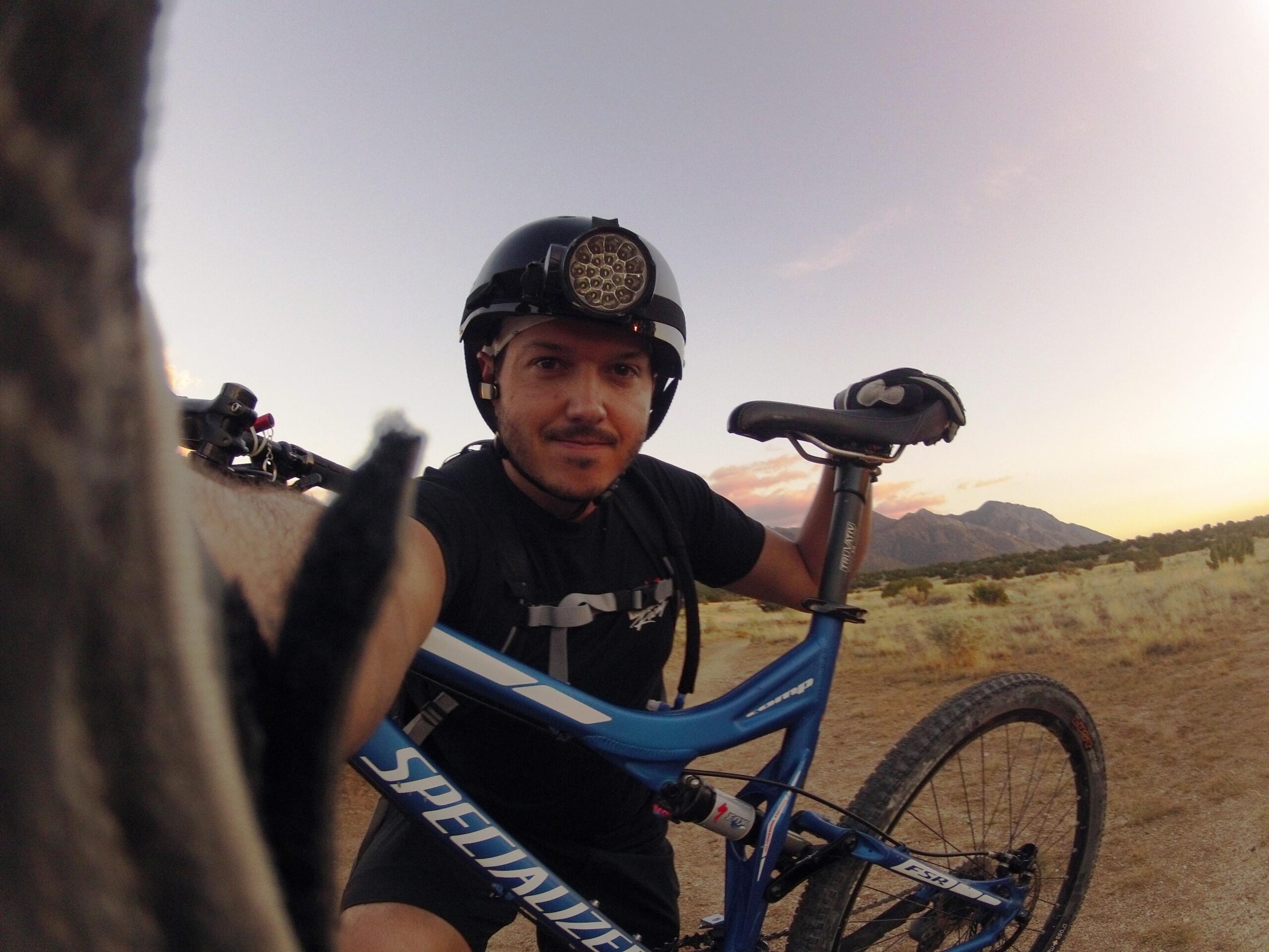 Specialized Stumpjumper FSR: A person wearing a helmet and biking gear takes a selfie while leaning on a blue mountain bike. The background features a scenic view of mountains and a sunset sky in a natural, outdoor setting.