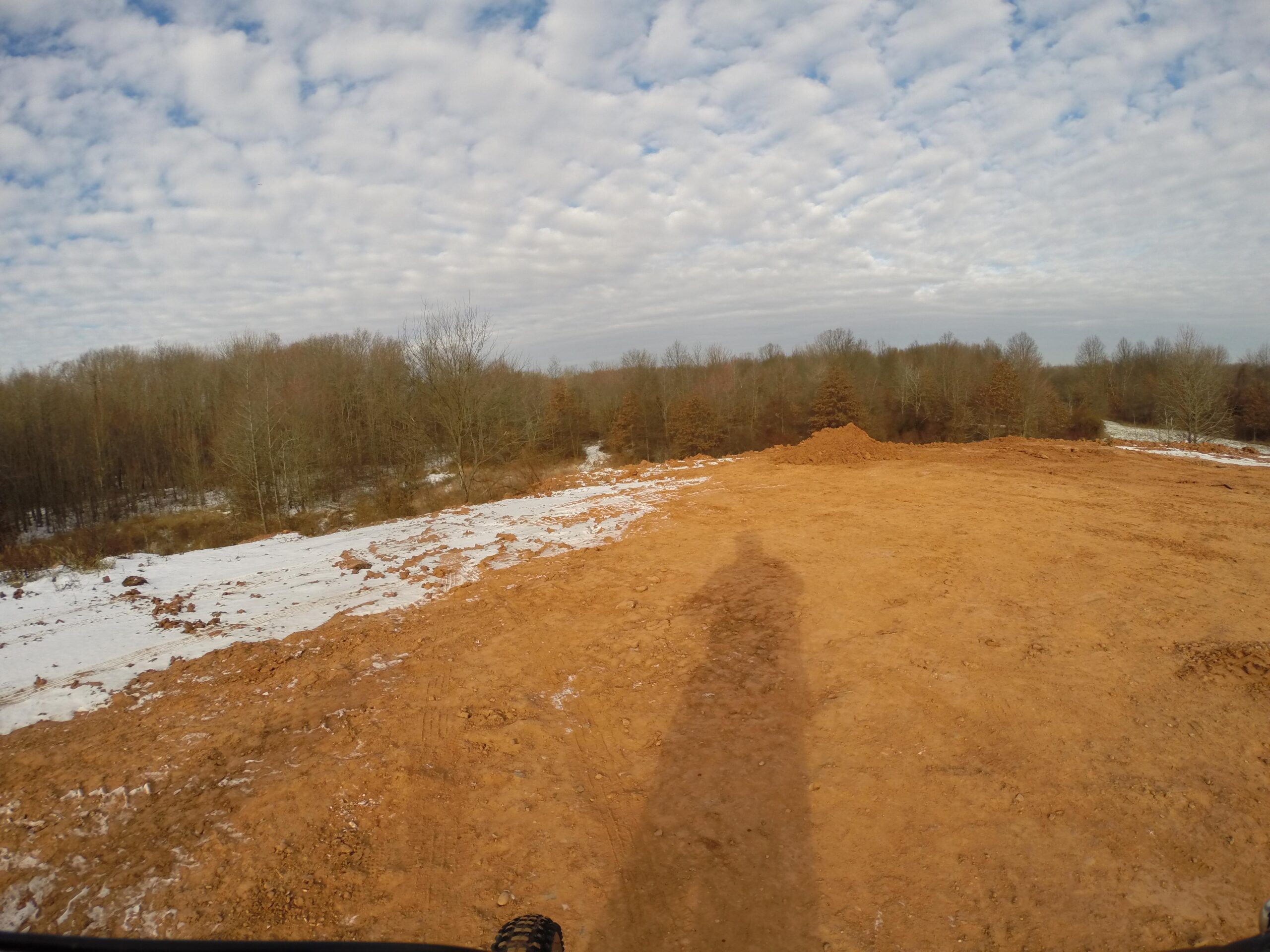 A panoramic view of a dirt landscape with patches of snow, featuring a cloudy sky. The foreground shows a sandy, uneven terrain, while the background consists of trees without leaves, indicating a winter setting. A shadow is visible in the bottom left corner, suggesting a person or object in the vicinity. Long Pond mountain bike trail.