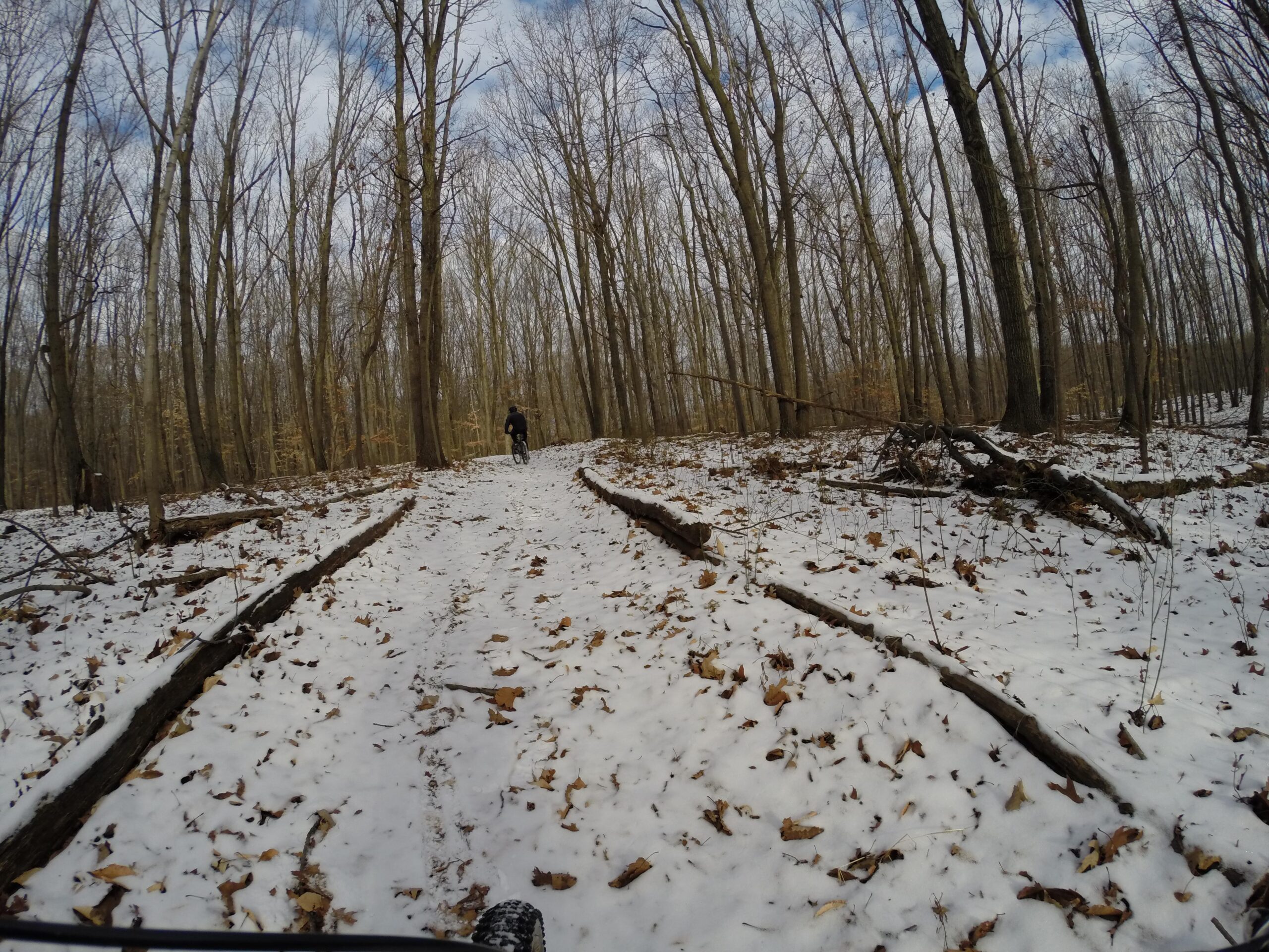 A cyclist riding a mountain bike on a snowy trail in a dense forest with bare trees and scattered fallen leaves. The scene features a mix of snow-covered ground and exposed earth, with a cloudy sky overhead. Long Pond mountain bike trail.