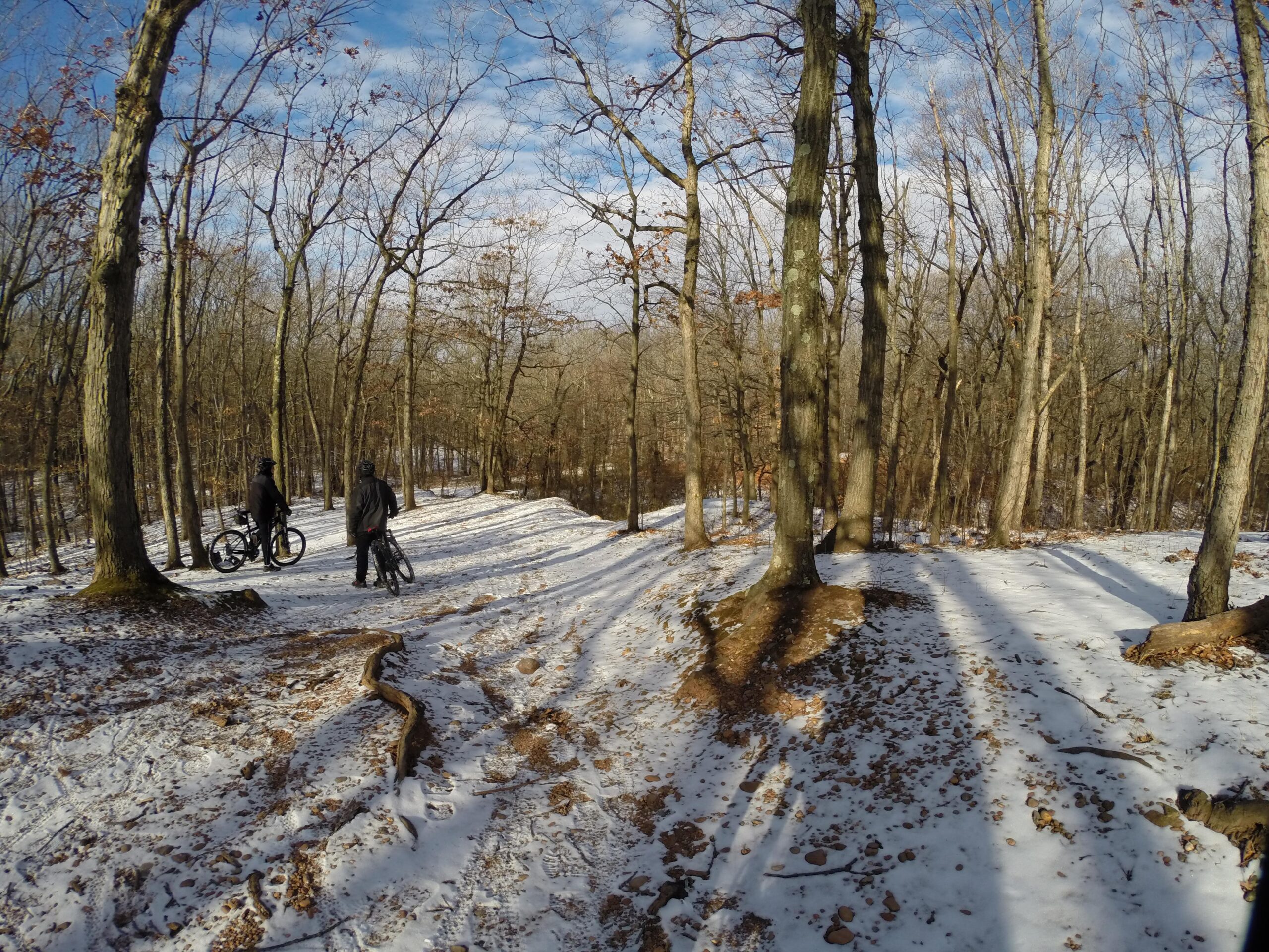 Two mountain bikers pause on a snowy trail in a forested area, surrounded by bare trees under a partly cloudy sky. The ground is covered in patches of snow and fallen leaves, and the sunlight casts long shadows on the trail. Long Pond mountain bike trail.