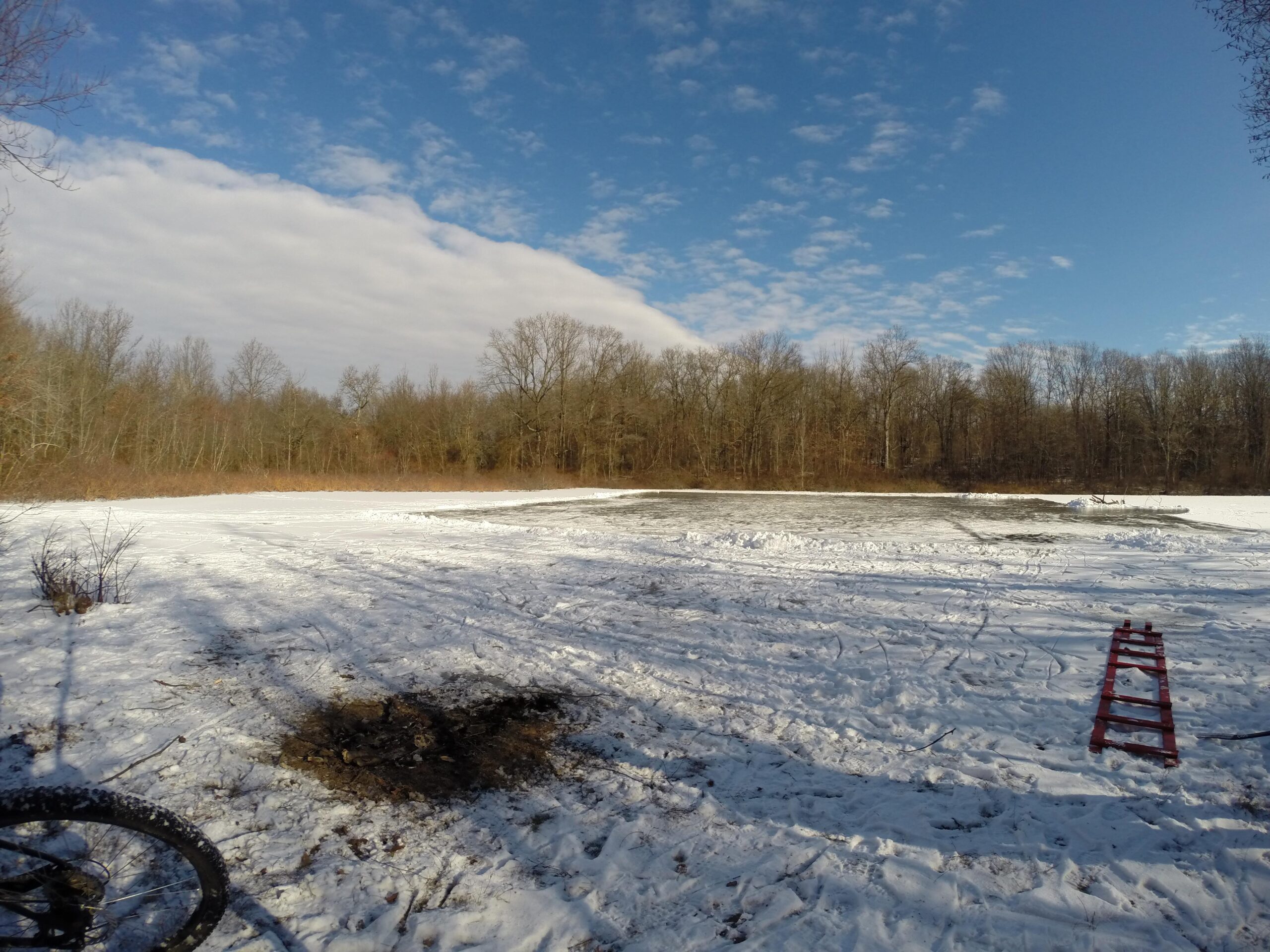 A snowy landscape with a mix of cleared ground and untouched snow, surrounded by barren trees in the background. The sky is partly cloudy with patches of blue. A red ladder rests on the snow, and a bicycle wheel is partially visible in the foreground. Long Pond mountain bike trail.