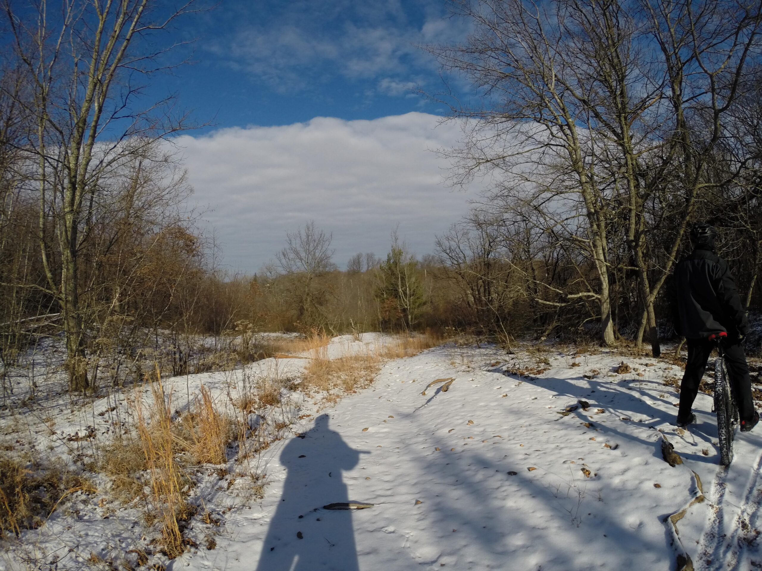 A winter landscape features a snowy path surrounded by leafless trees under a blue sky with clouds. A person in winter clothing is standing on the path, casting a long shadow on the snow. The scene captures the tranquility of nature in a rural setting during winter. Long Pond mountain bike trail.