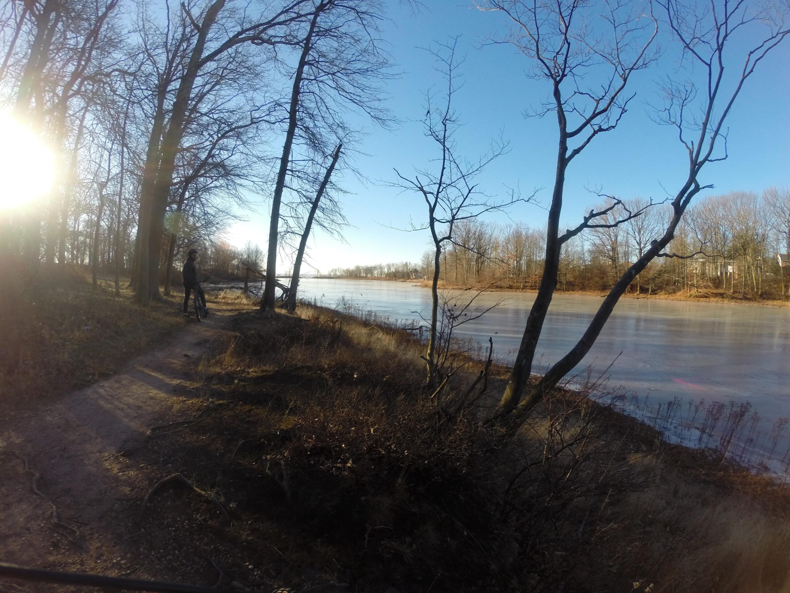 A serene winter landscape featuring a partially frozen lake bordered by leafless trees. A person stands by the water's edge with a bicycle, basking in the warm sunlight. The clear blue sky contrasts with the earthy tones of the surroundings, creating a peaceful outdoor scene. Wolfes Pond park mountain bike trail.