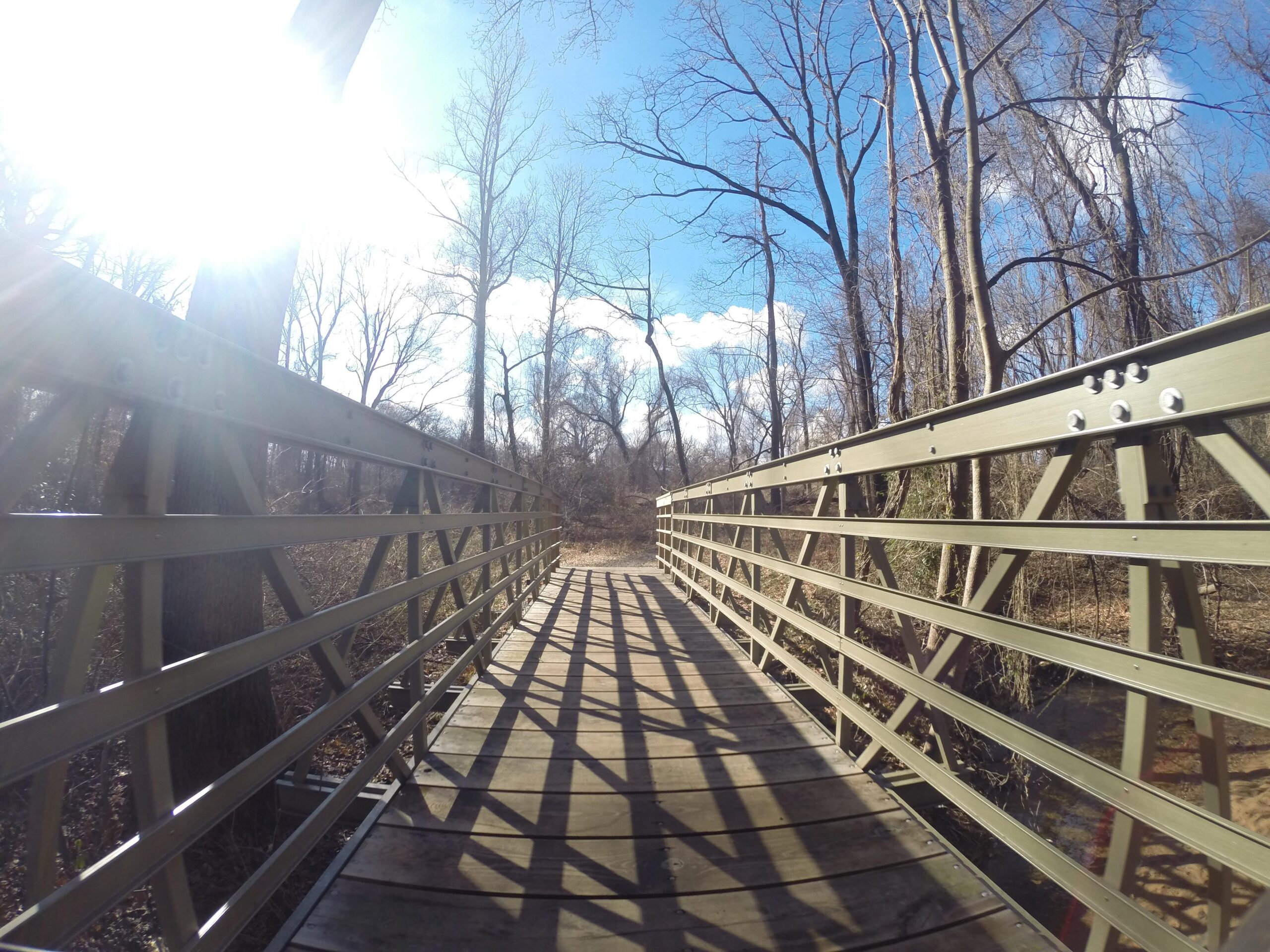 A wooden footbridge crosses a tranquil scene in a wooded area. Sunlight filters through the trees, casting shadows on the bridge’s surface. The sky is partly cloudy, and bare trees line the path, suggesting a late fall or early spring setting. Huber Woods mountain bike trail.