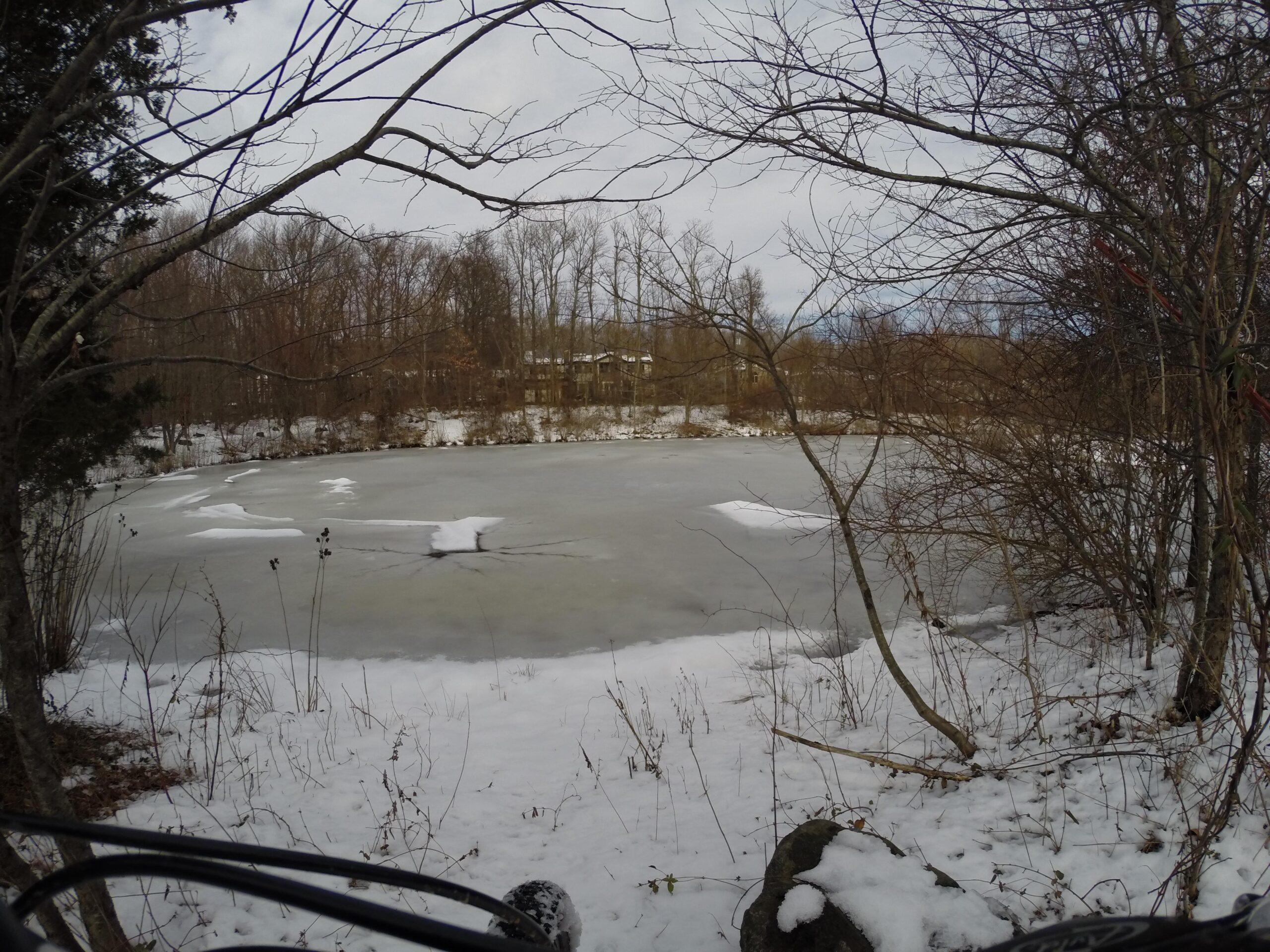A partially frozen pond surrounded by bare trees and snow-covered ground. Ice patches float on the water's surface, with a cloudy sky in the background. The scene conveys a serene winter landscape. Trails seperated by streets mountain bike trail.