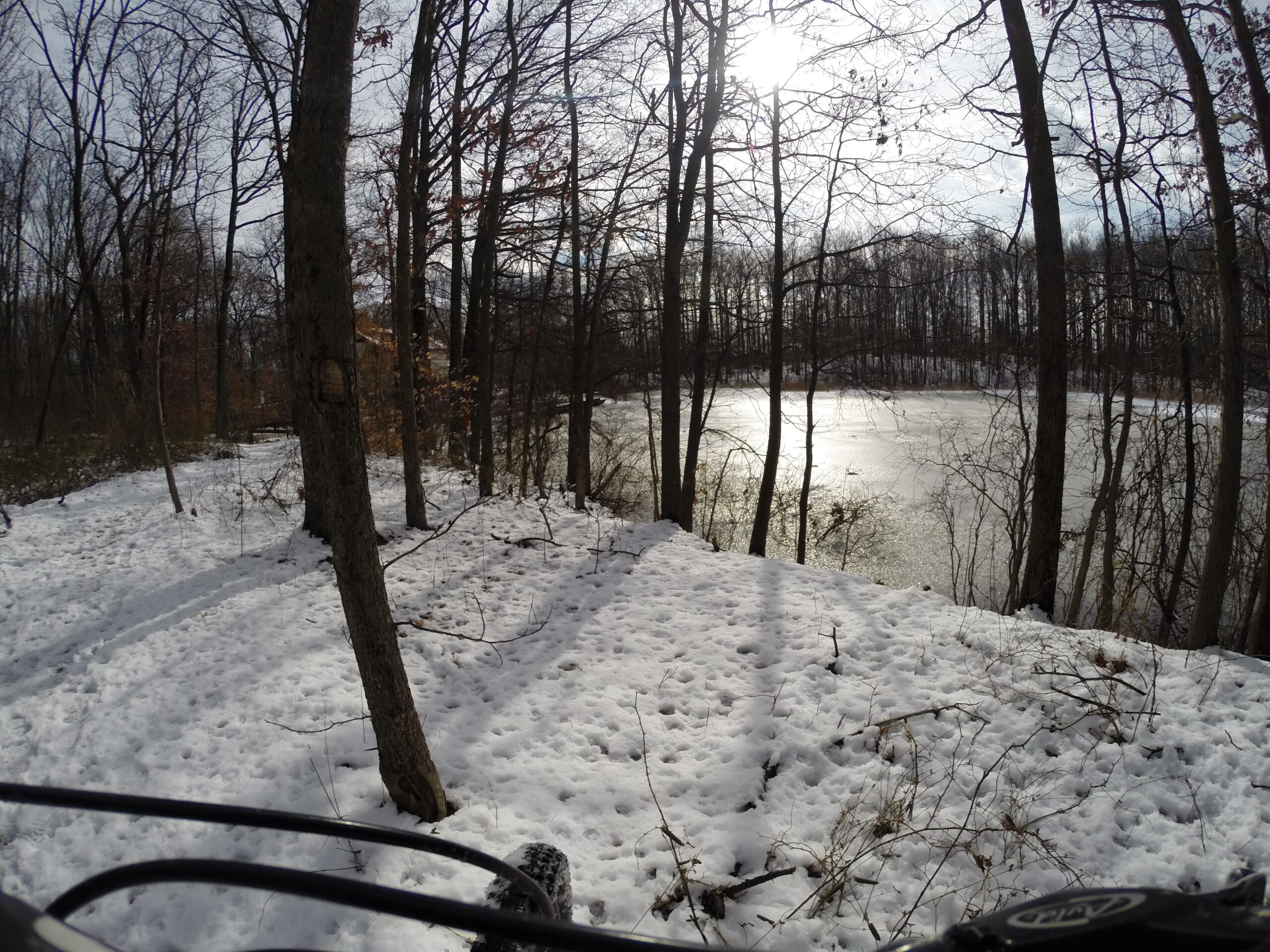 A snowy forest scene with bare trees lining the edge of a partially frozen pond. The sun is shining through the clouds, casting shadows on the snow-covered ground. A bicycle handlebar is visible in the foreground, suggesting an outdoor biking adventure in winter. Trails seperated by streets mountain bike trail.