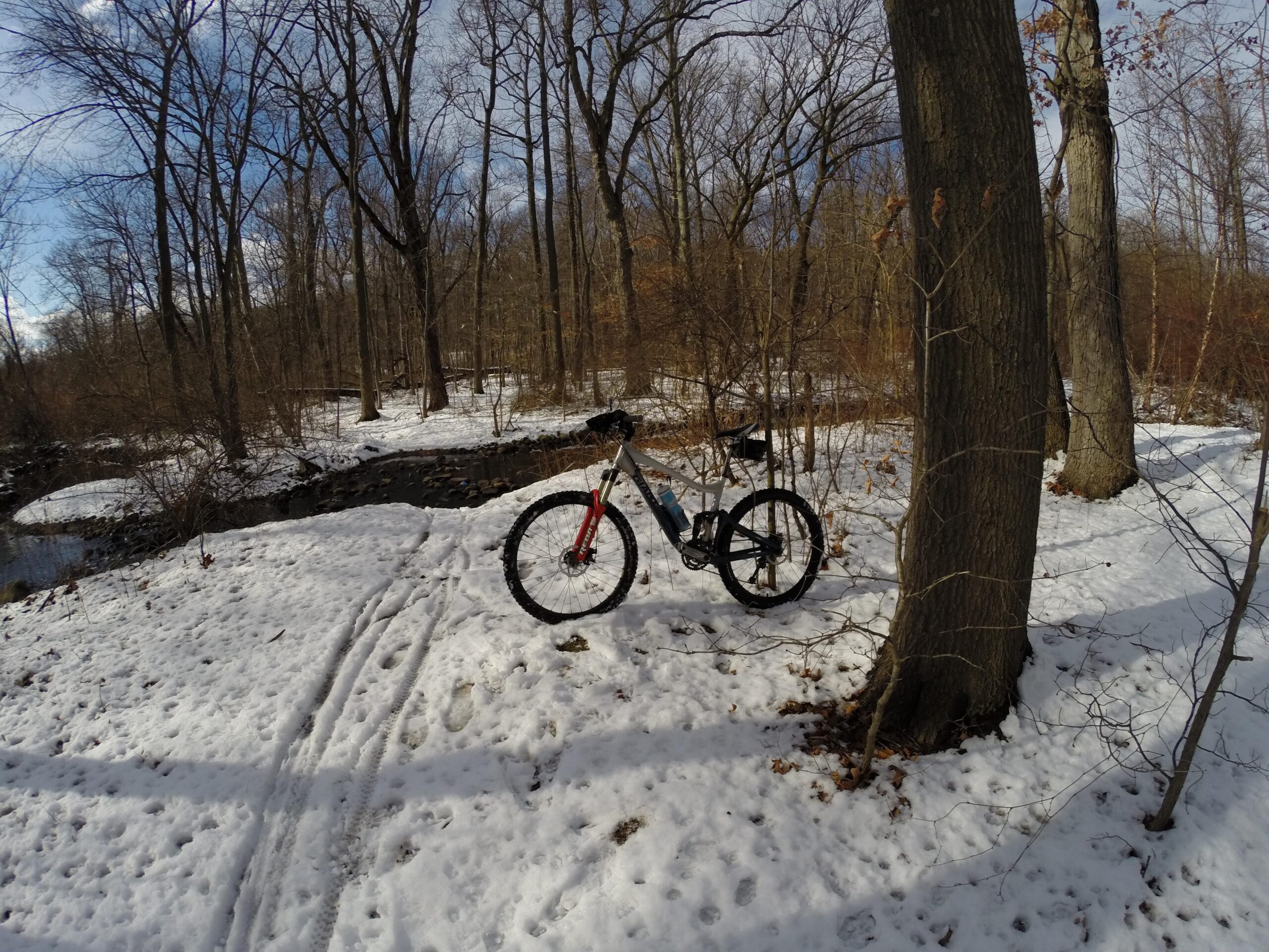 A mountain bike resting on snowy ground next to a frozen creek, surrounded by bare trees in a winter landscape with patches of blue sky. Wolfes Pond park mountain bike trail.