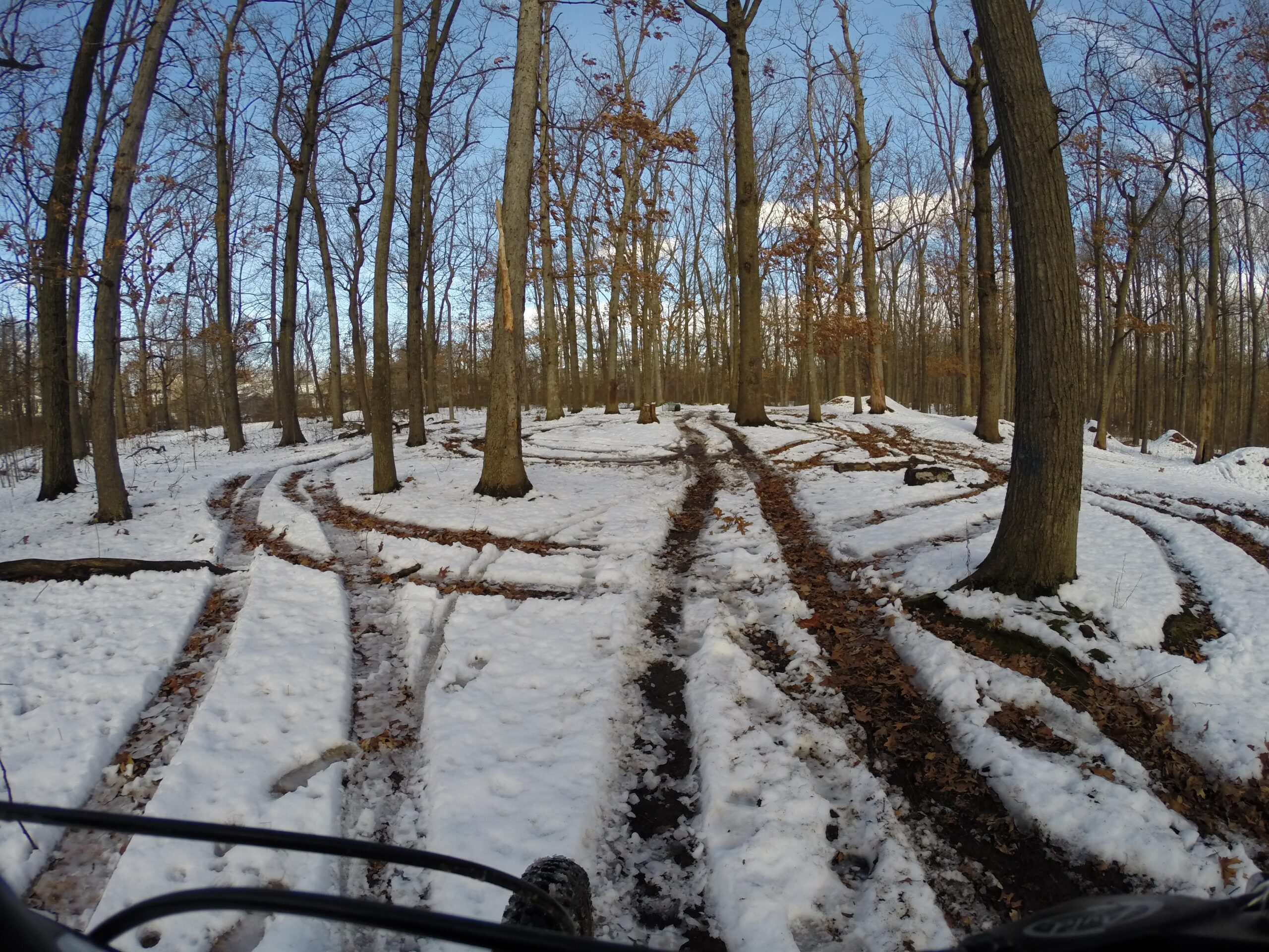 A snow-covered trail winding through a forest of bare trees, with tracks left by bicycle tires and fallen leaves scattered on the ground. The scene is illuminated by daylight, showcasing a clear blue sky. Wolfes Pond park mountain bike trail.