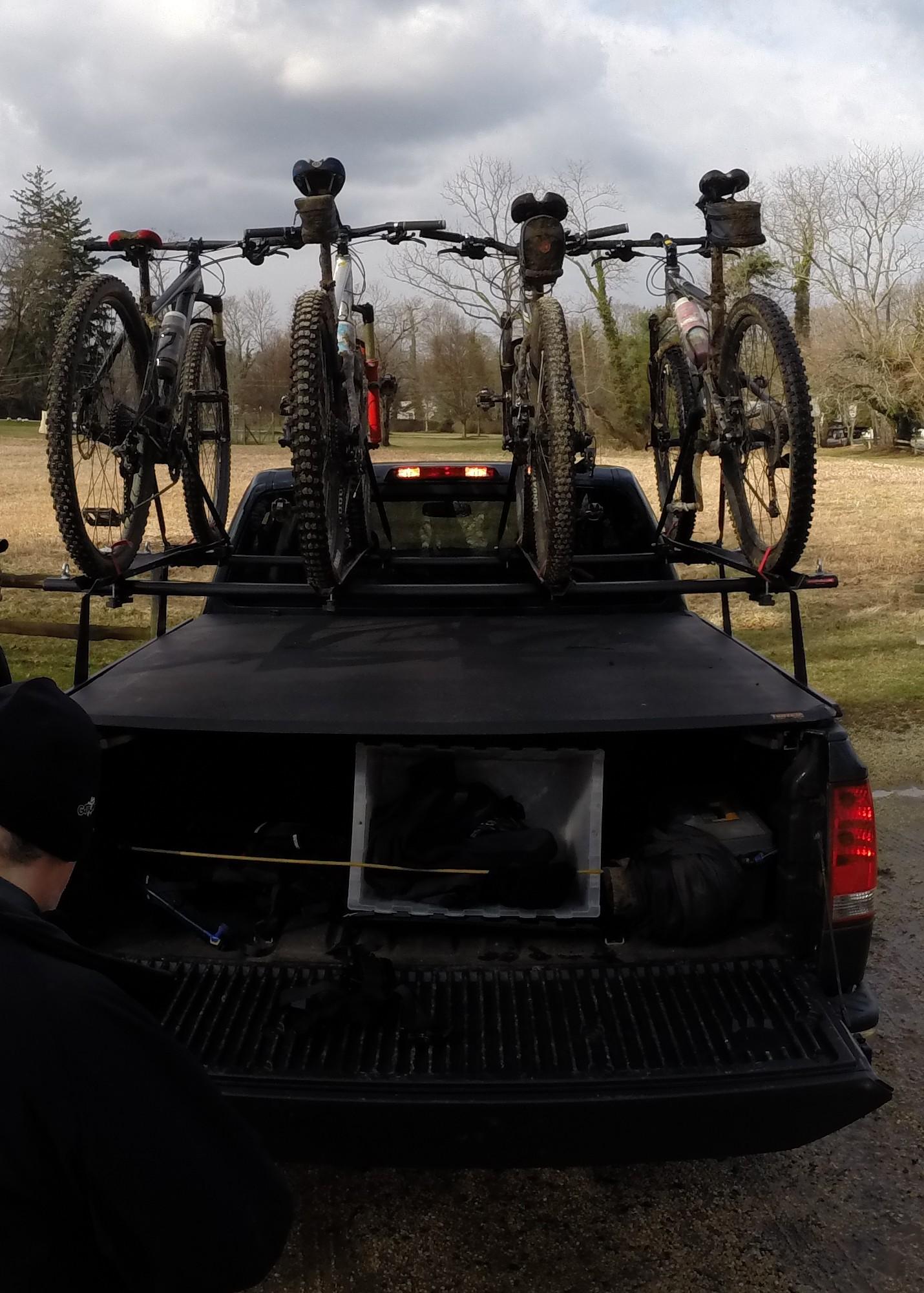 A pickup truck with four mountain bikes securely mounted on a bike rack. The bikes are dirty, indicating recent use, and there is an open truck bed revealing various gear and equipment. In the foreground, a person wearing a black jacket and hat is seen near the truck. The background features a rural landscape with bare trees and cloudy skies. Huber Woods mountain bike trail.