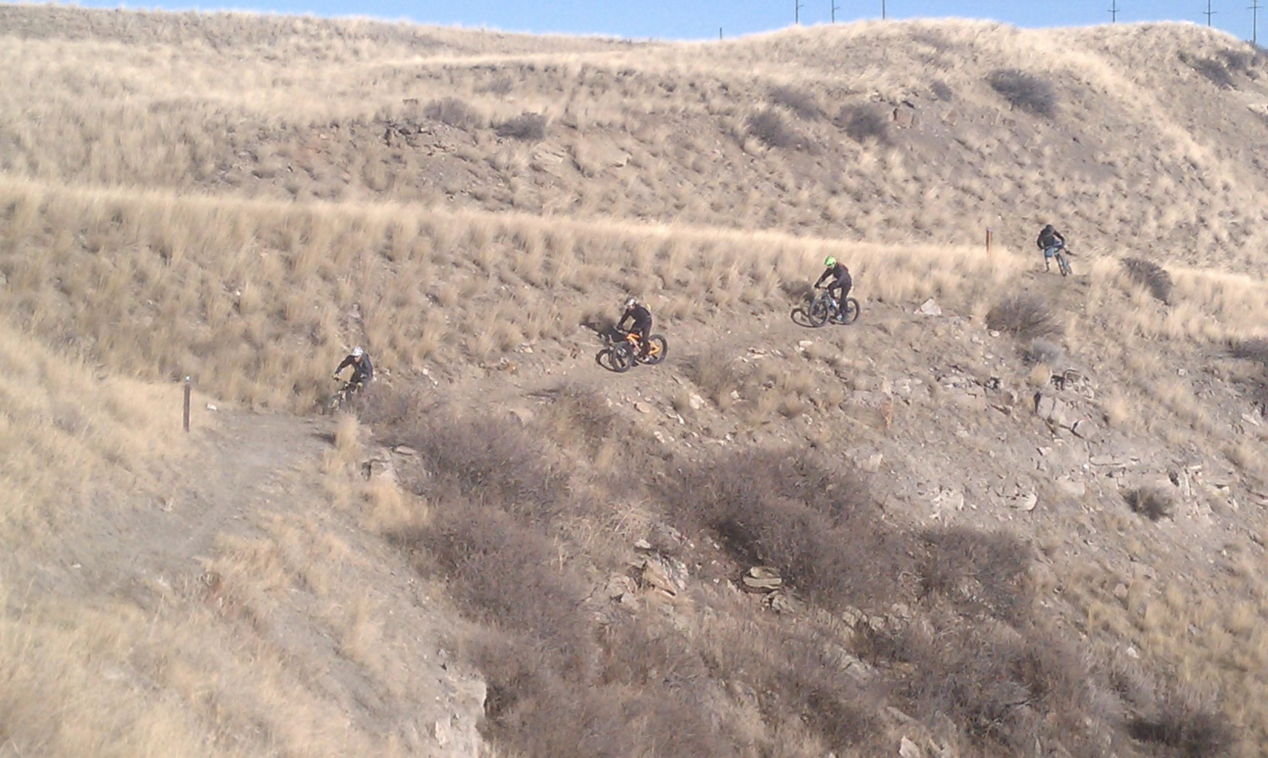 A group of mountain bikers navigating a rugged trail on a grassy hillside, with scattered rocks and sparse vegetation surrounding them. The background features rolling hills under a clear blue sky. New Video Game/Yeah Kid mountain bike trail.