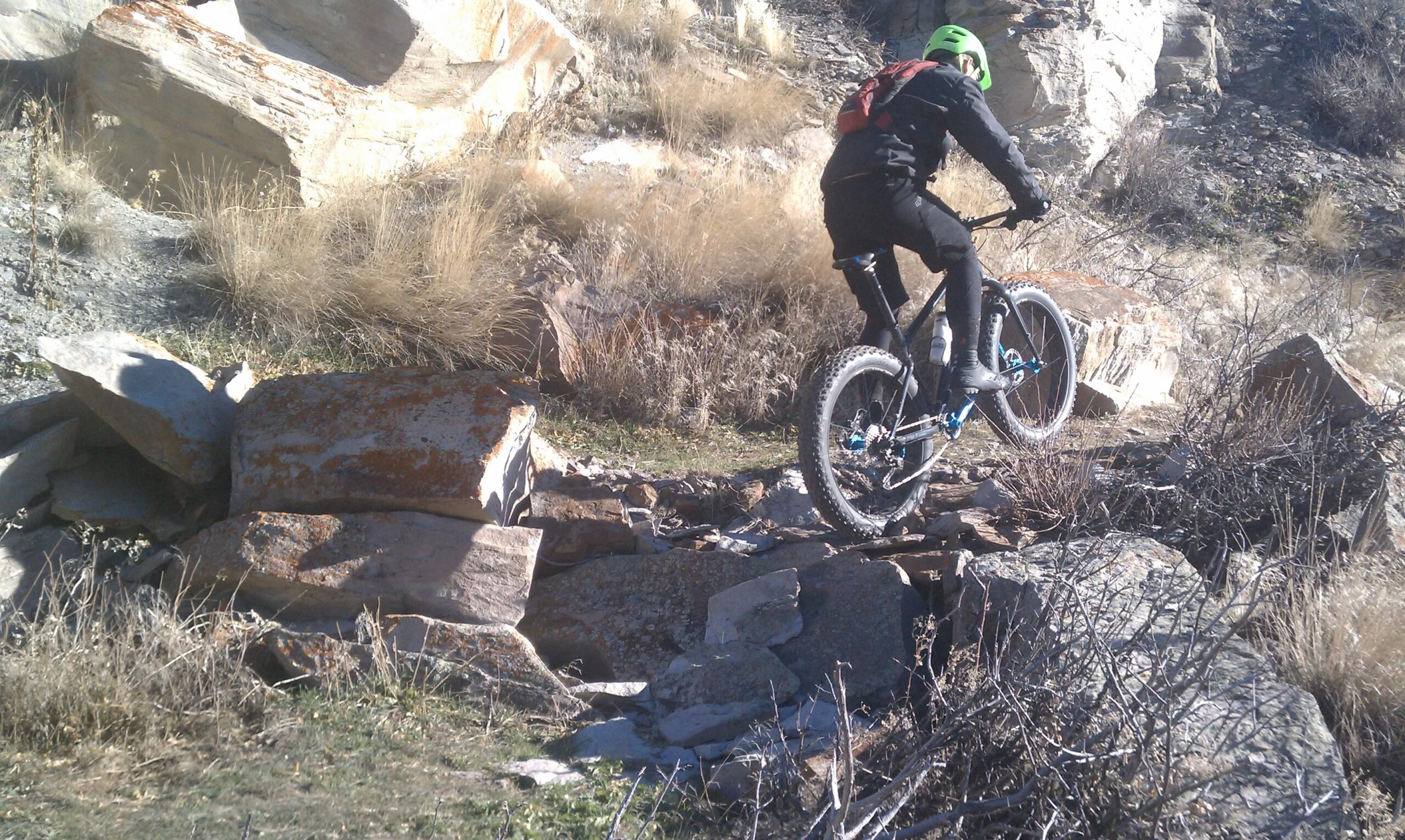 A mountain biker navigating rocky terrain. The rider is wearing a green helmet and black gear, showcasing a dynamic pose as they maneuver over large stones in an outdoor setting. Dry grass and sparse vegetation surround the area, emphasizing the rugged landscape. New Video Game/Yeah Kid mountain bike trail.