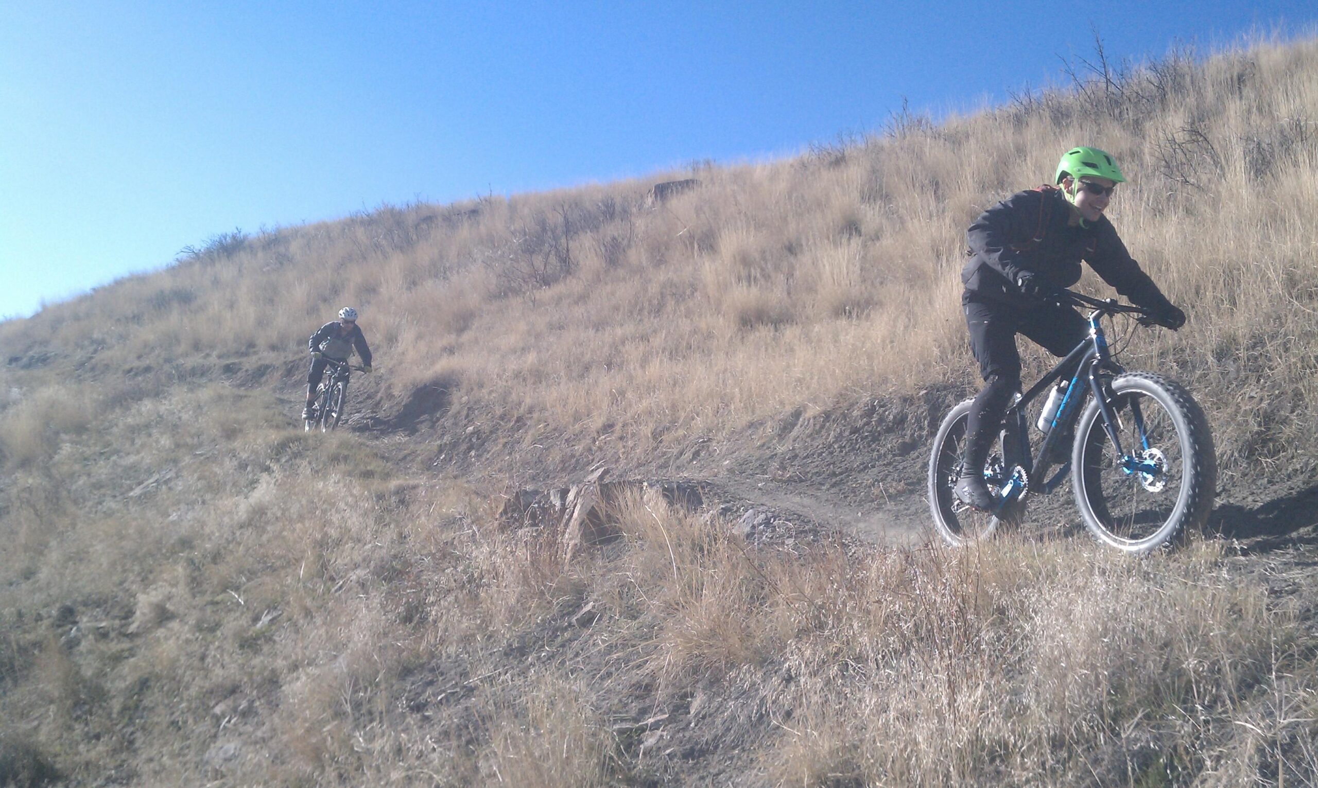 Two mountain bikers riding along a dirt trail on a grassy hillside under a clear blue sky. One biker is ascending, while the other is descending, both wearing helmets and appropriate cycling gear. New Video Game/Yeah Kid mountain bike trail.