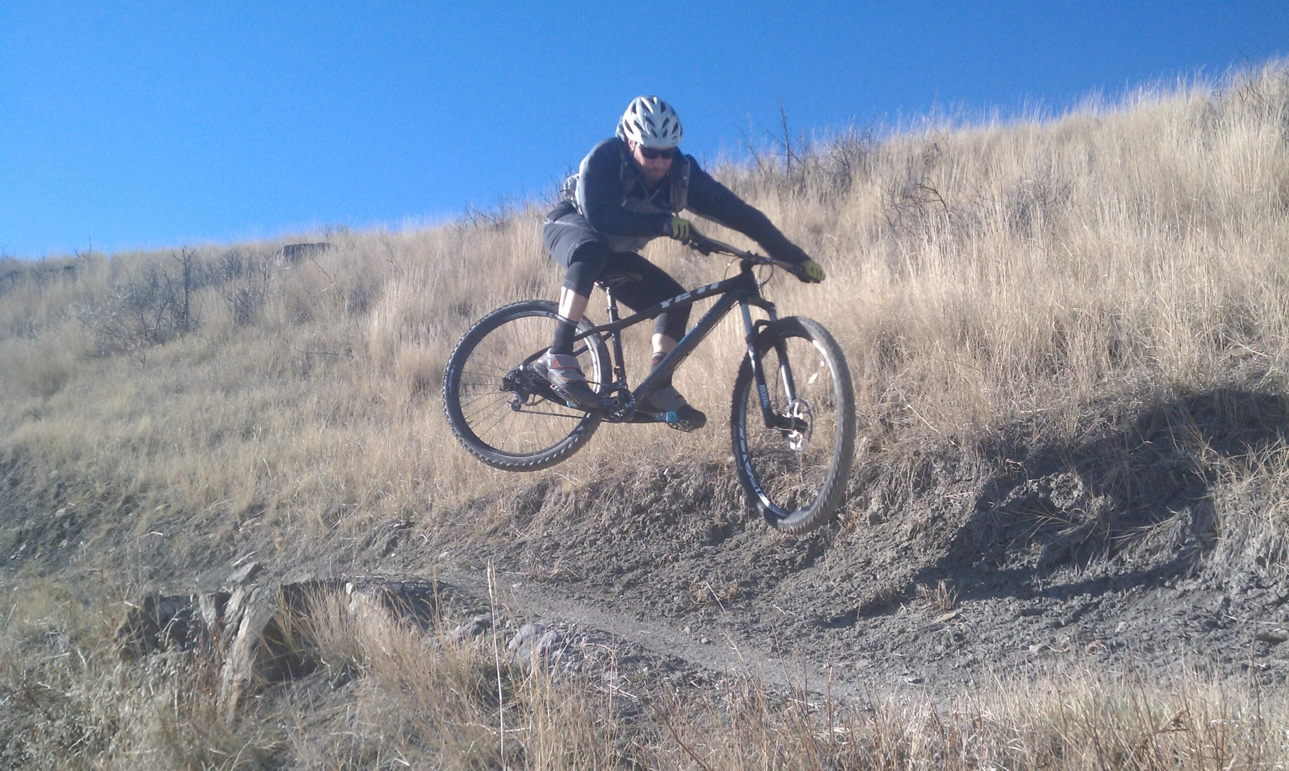 A cyclist wearing a helmet and sunglasses performs a jump on a mountain bike over a dirt trail, surrounded by dry grass and a clear blue sky. New Video Game/Yeah Kid mountain bike trail.