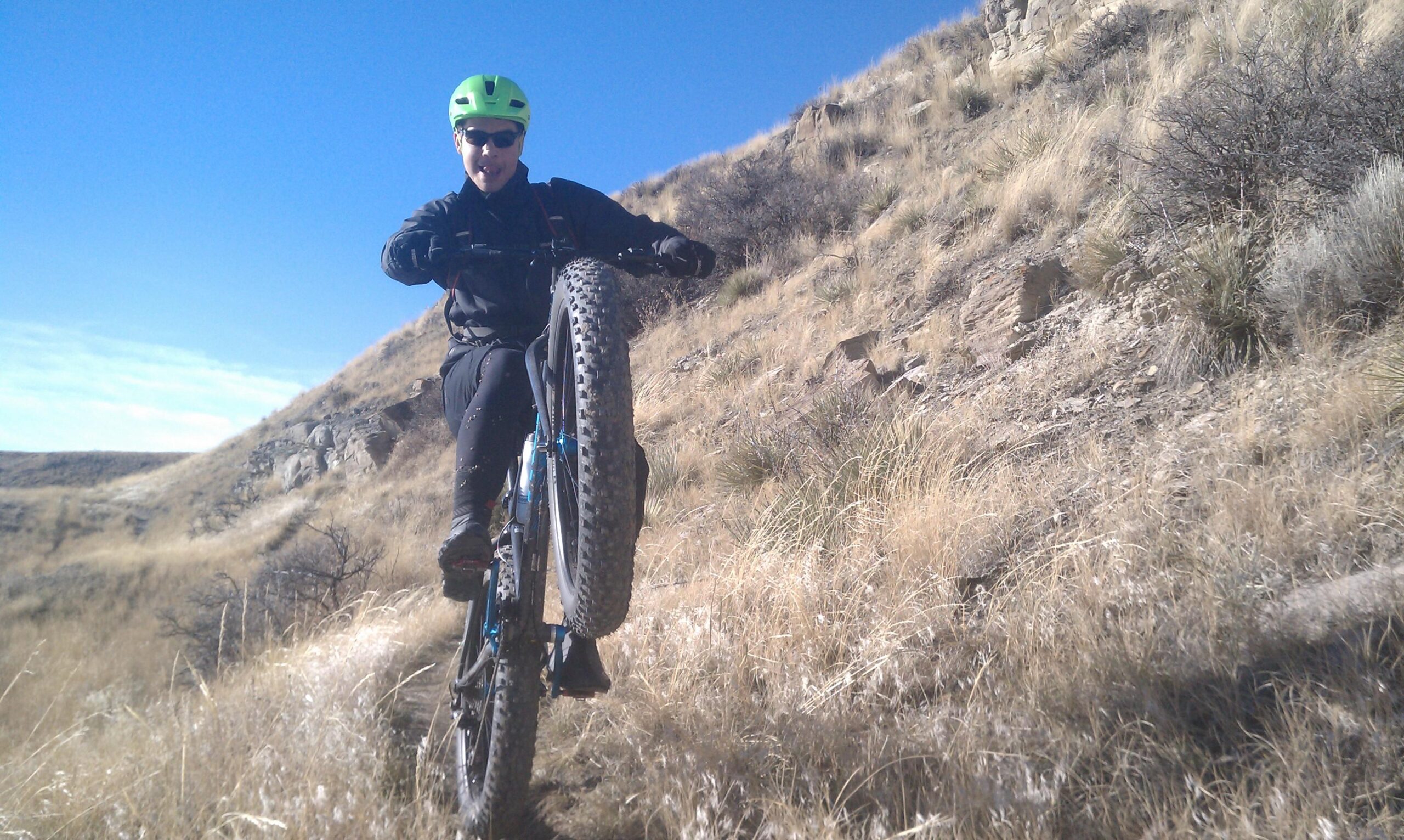 A person riding a mountain bike on a rugged trail, lifting the front wheel off the ground. The rider is wearing a bright green helmet and sunglasses, with a background of dry grass and rocky terrain under a clear blue sky. New Video Game/Yeah Kid mountain bike trail.