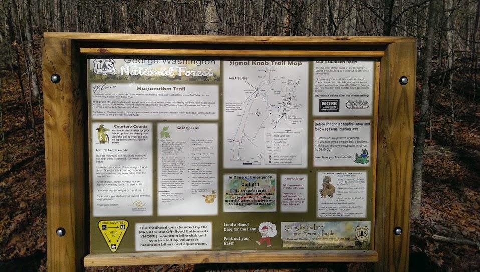 A wooden information board in a forest setting, displaying details about the Massanutten Trail and Signal Knob Trail map. The sign includes safety tips, emergency contact information, and guidelines for trail courtesy, with text and illustrations. Surrounding trees are visible in the background. Elizabeth Furnace mountain bike trail.