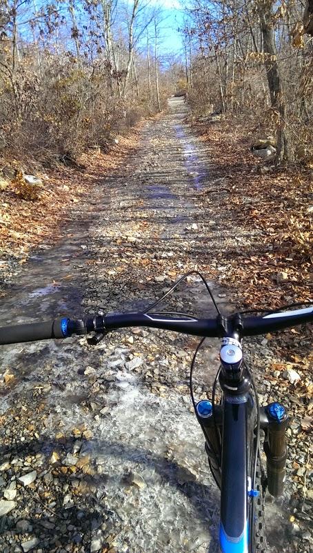 A mountain bike is shown from the rider's perspective, positioned on a rocky trail surrounded by trees. The path is covered in fallen leaves and patches of ice, leading into a sunny, clear blue sky in the distance. Elizabeth Furnace mountain bike trail.