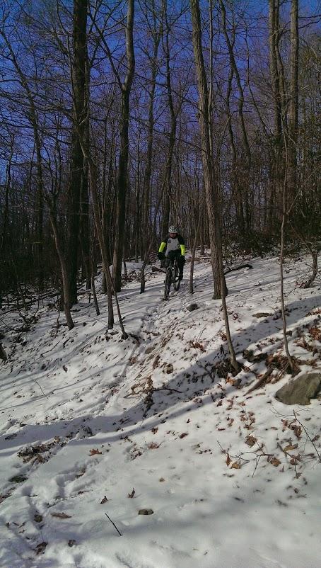 A mountain biker navigates a snow-covered trail in a forest, surrounded by bare trees under a clear blue sky. The rider, wearing bright yellow sleeves for visibility, is maneuvering up a slight incline on the path. Frederick Watershed mountain bike trail.