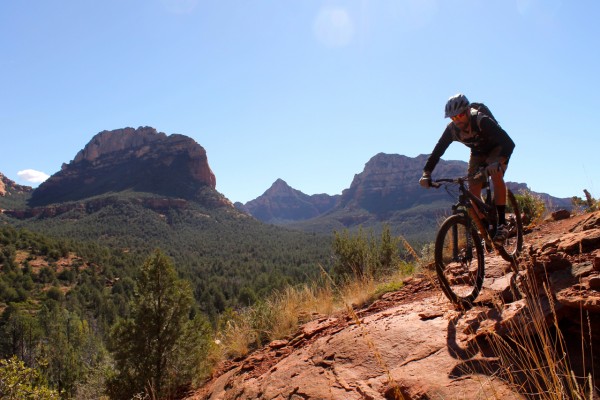 A mountain biker navigating a rocky trail with rugged mountains and a clear blue sky in the background. Lush greenery surrounds the trail, highlighting a scenic outdoor environment conducive to biking.
