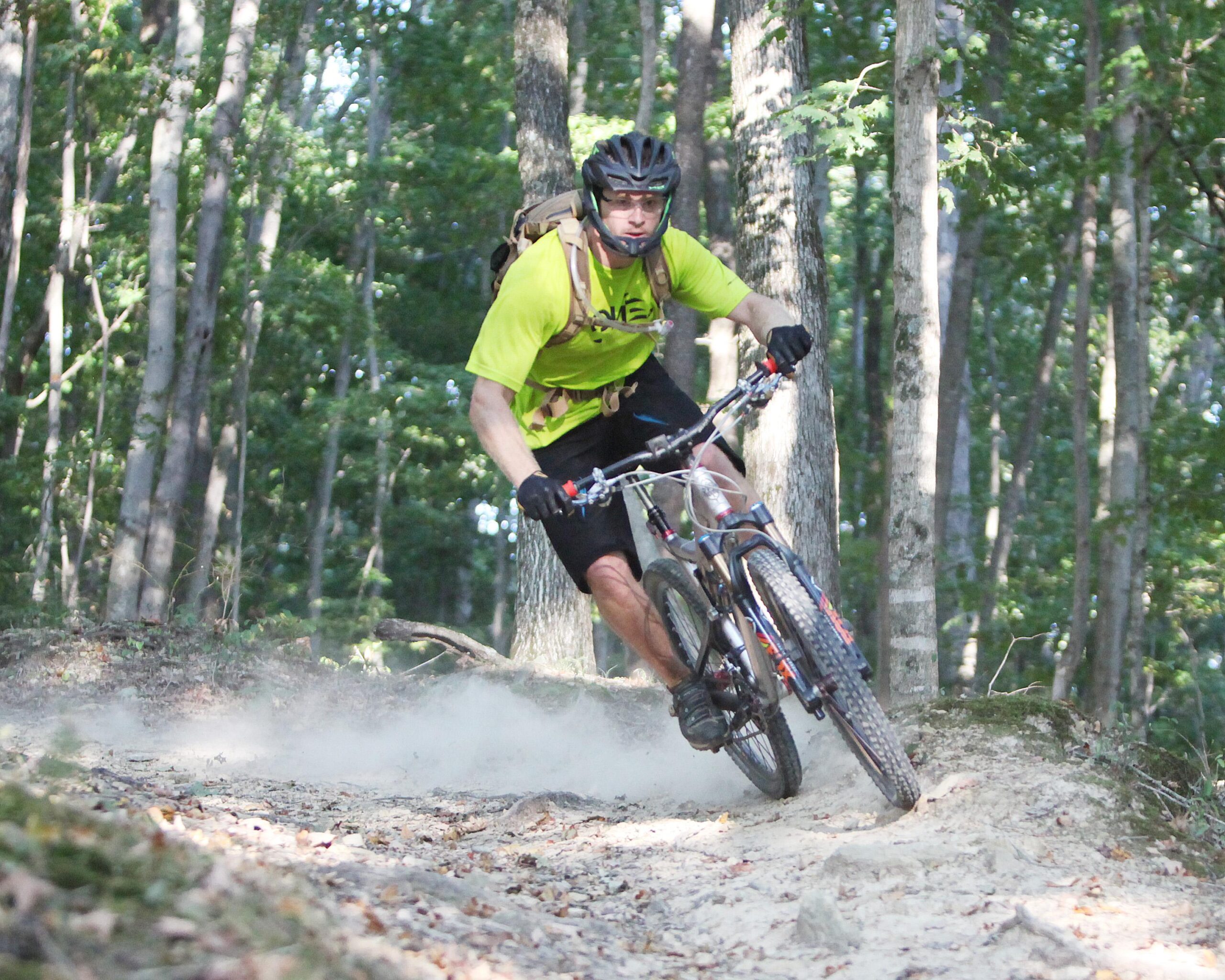 A mountain biker in a bright yellow shirt and black shorts rides a mountain bike on a dirt trail through a wooded area. Dust is kicked up behind the bike as the rider leans into a turn, surrounded by tall trees and greenery. Green River mountain bike trail.