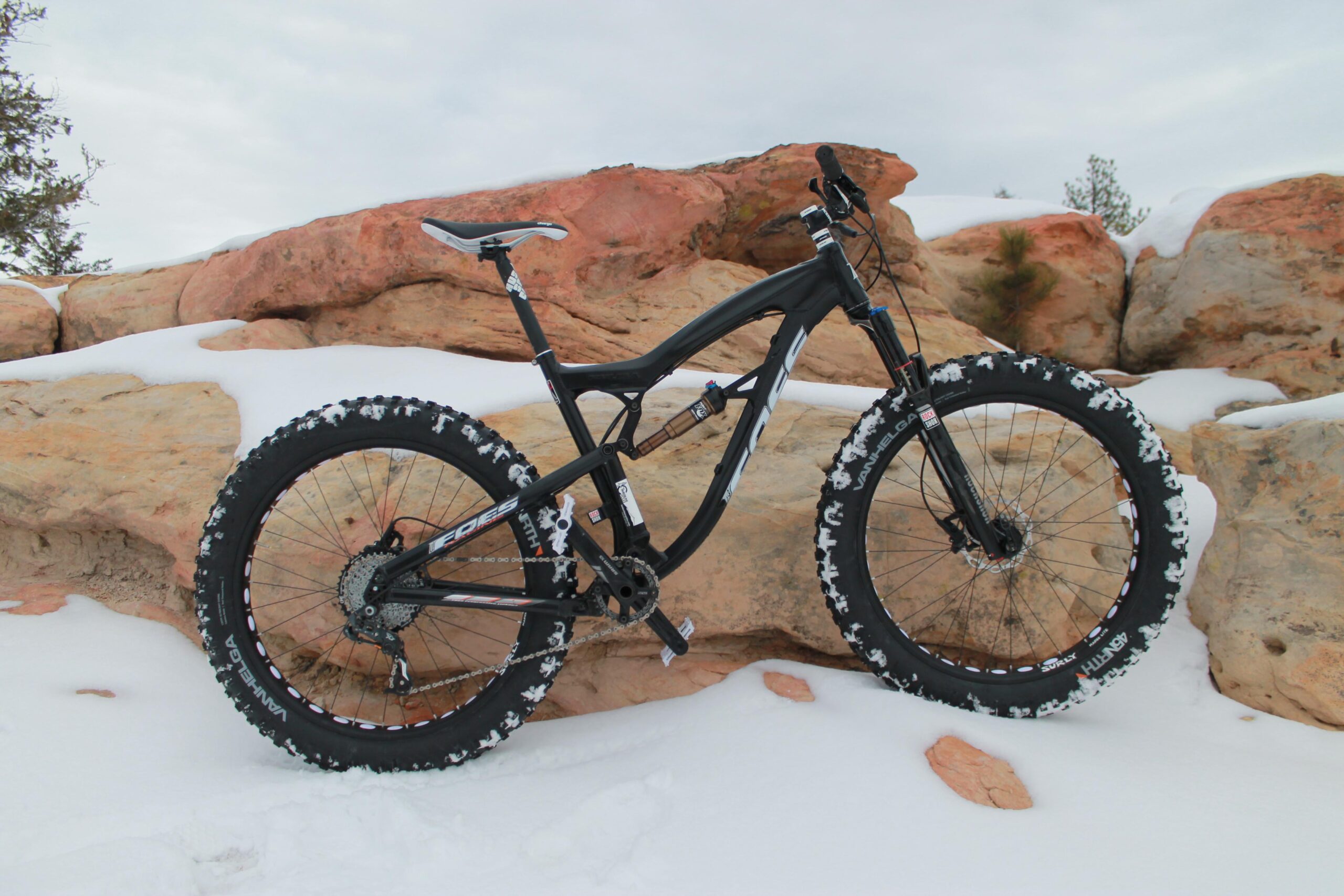 Foes Mutz: A black fat tire bike parked on snow-covered ground next to rocky terrain. The bike features wide tires with some snow accumulation and is positioned against a backdrop of orange and gray rocks under a cloudy sky.