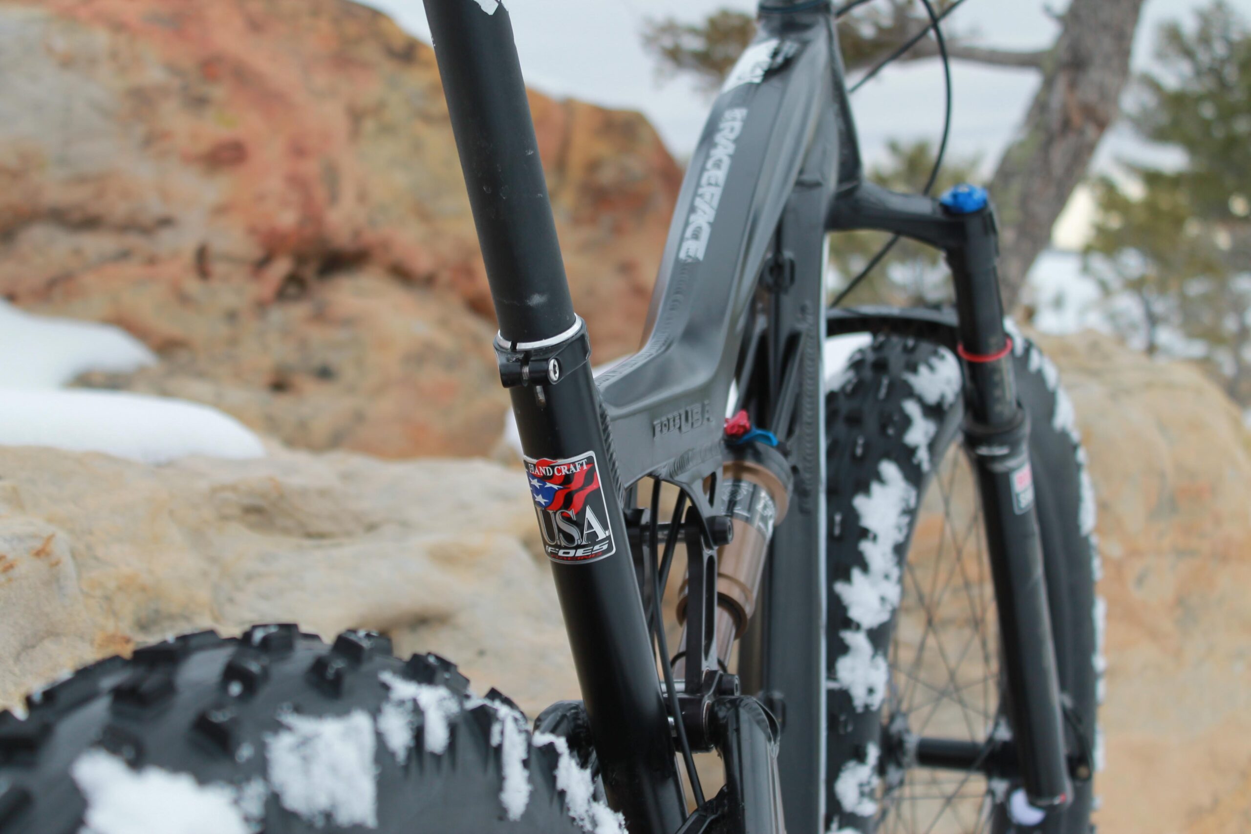 Foes Mutz: Close-up view of a fat bike frame showcasing its suspension components and a USA flag sticker. The background features rocky terrain with some snow, indicating a cold outdoor setting.