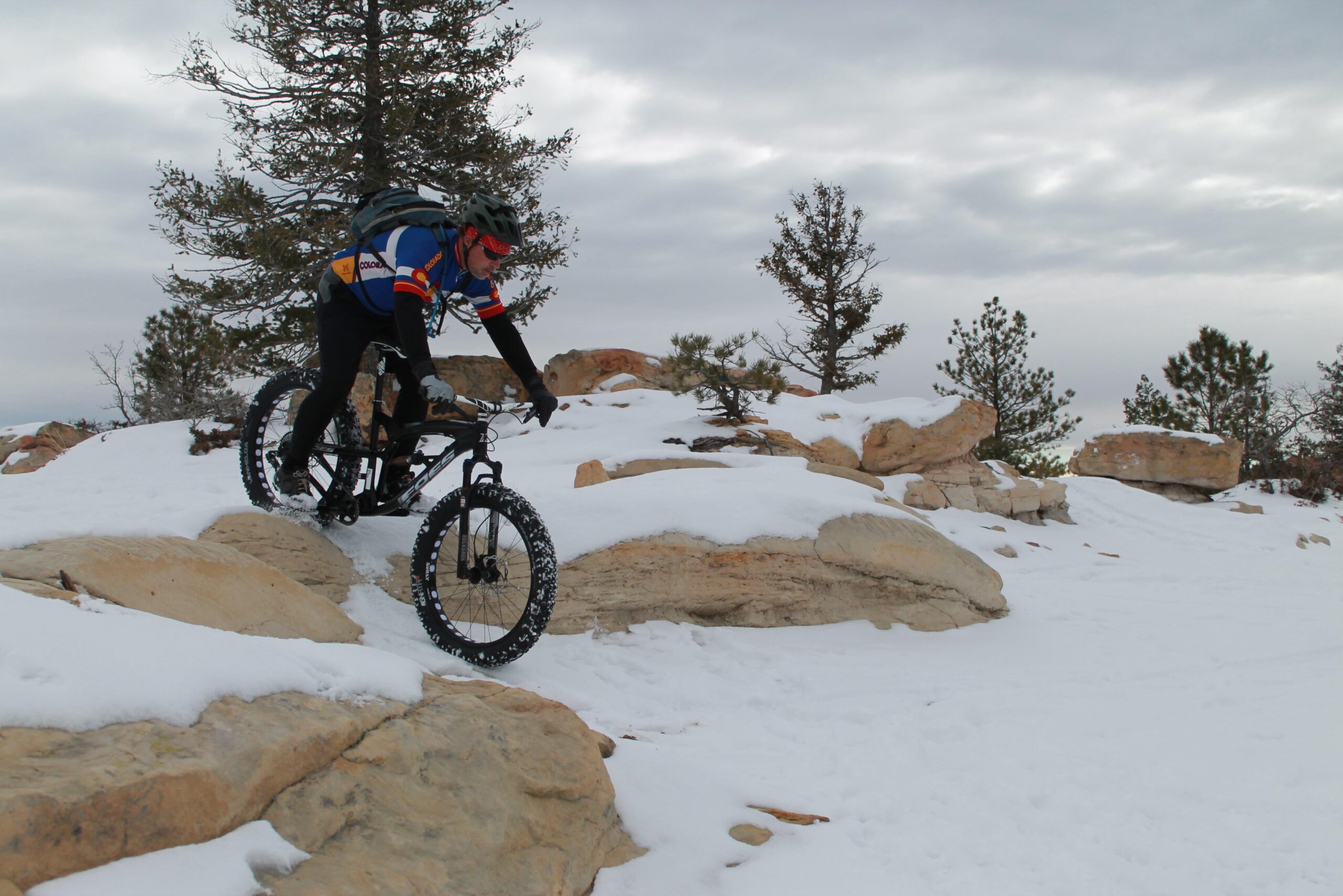 Foes Mutz: A mountain biker navigating rocky terrain while riding on snow-covered ground. The cyclist is wearing a helmet and winter gear, and a backpack, with evergreen trees in the background under a cloudy sky.