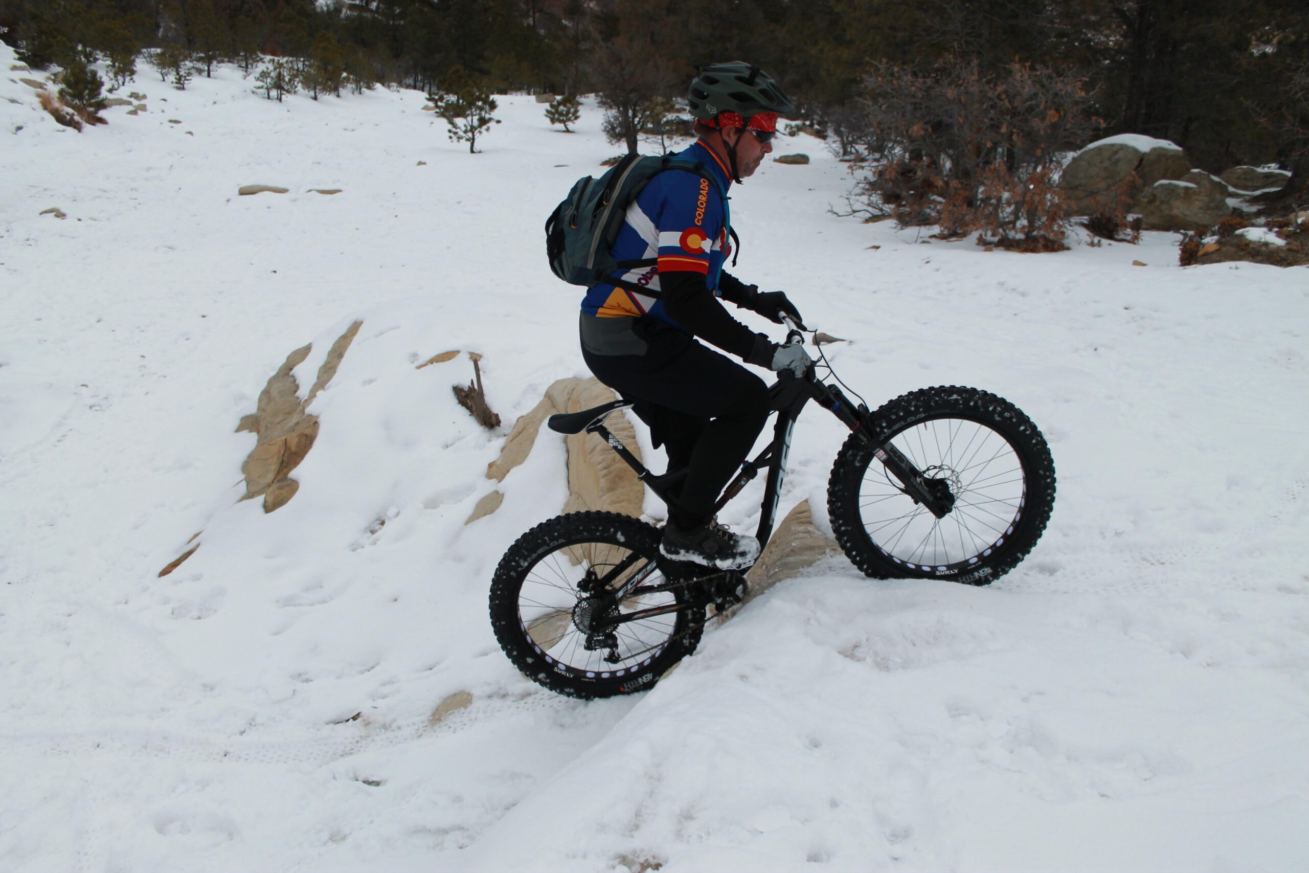 Foes Mutz: A cyclist riding a fat bike over a snow-covered trail, with snow-dusted rocks and pine trees in the background. The rider is wearing a helmet, sunglasses, and a colorful jersey, showcasing their skill as they navigate the terrain.