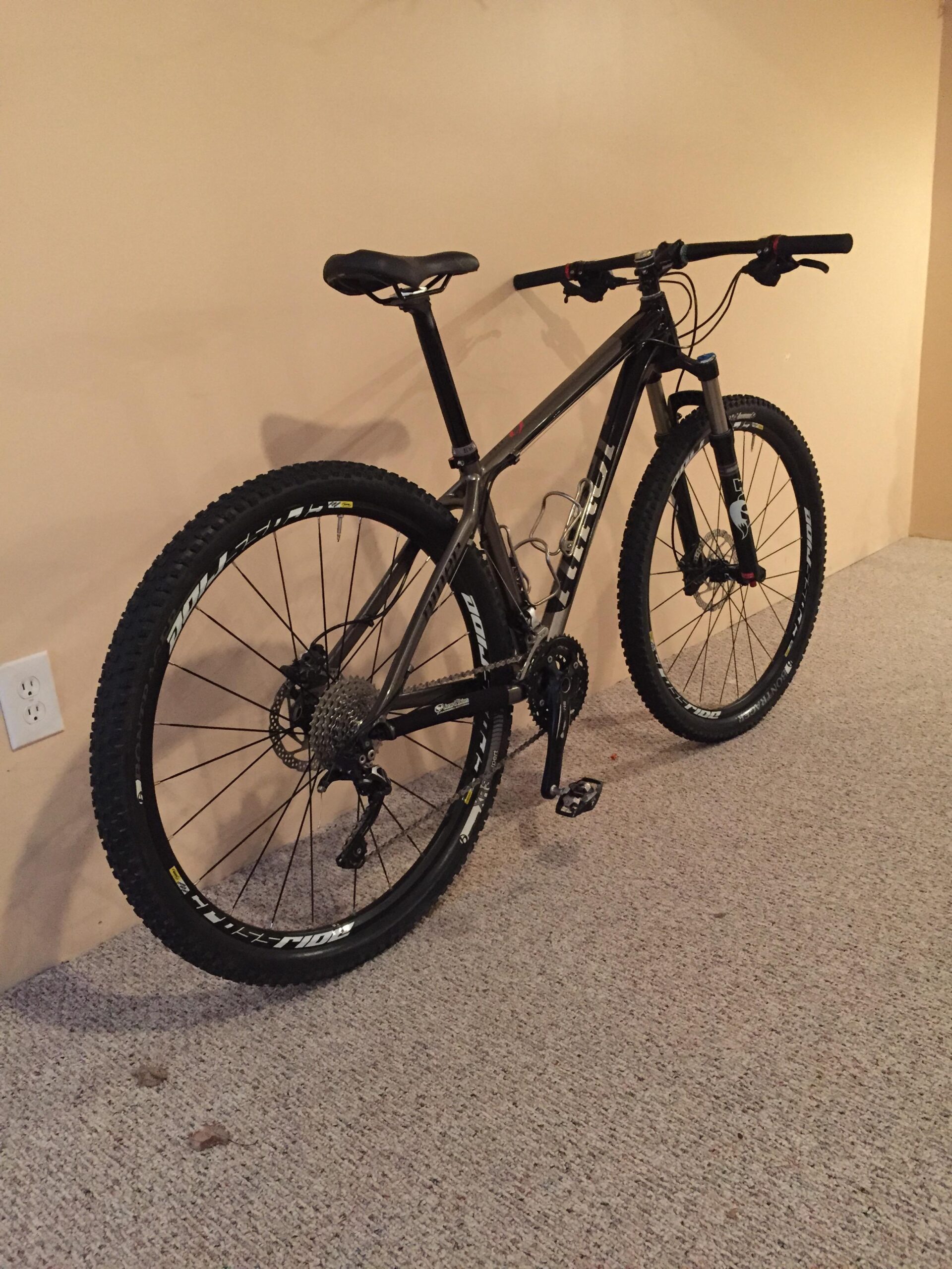 Niner Air 9: Mountain bike leaning against a beige wall, featuring large knobby tires, a sturdy frame, and a black seat. The bike is positioned on a carpeted floor with visible texture.