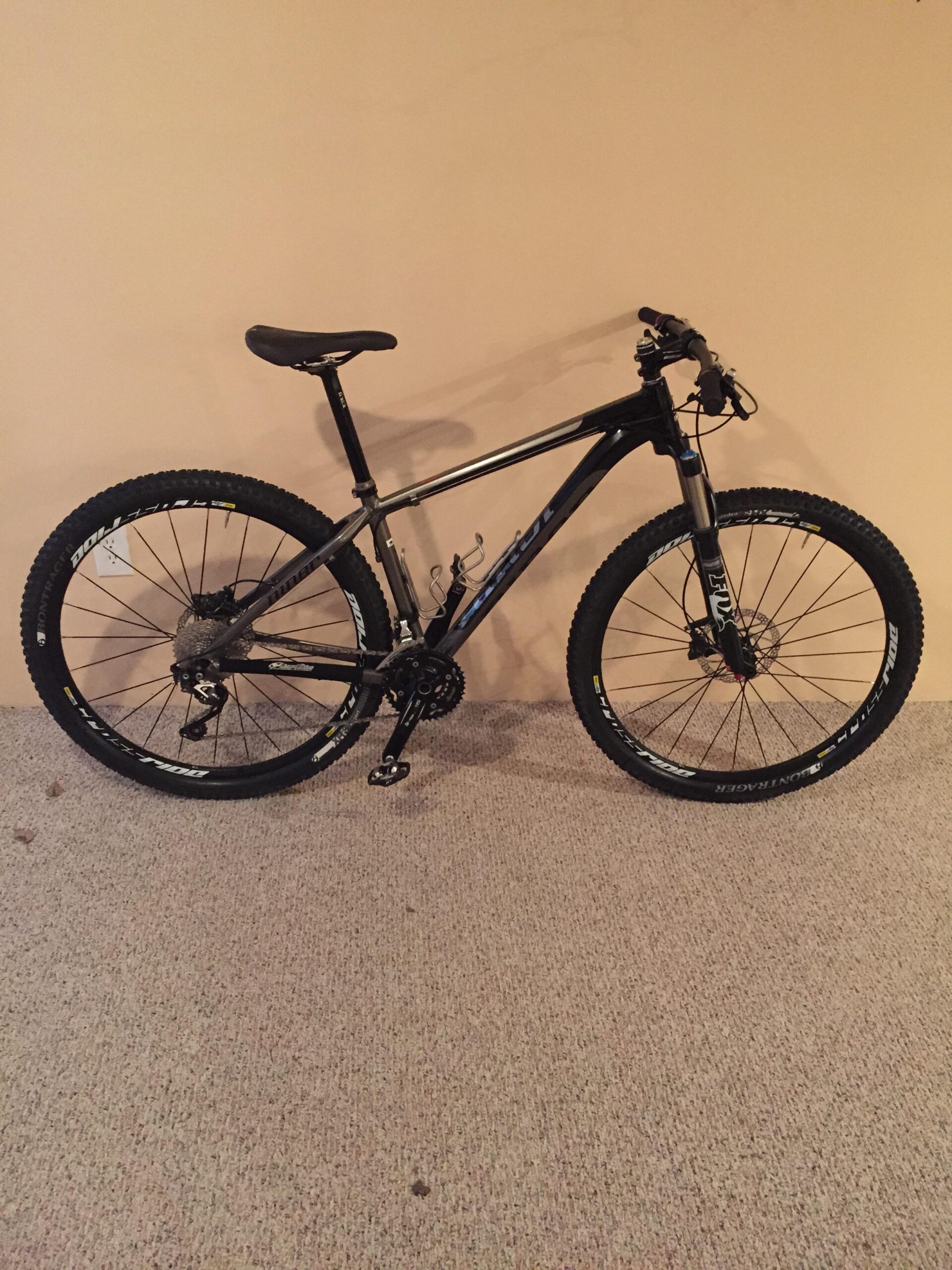 Niner Air 9: A black mountain bike leaning against a beige wall, featuring wide tires, front suspension, and a basic gear shifter, positioned on a carpeted surface.