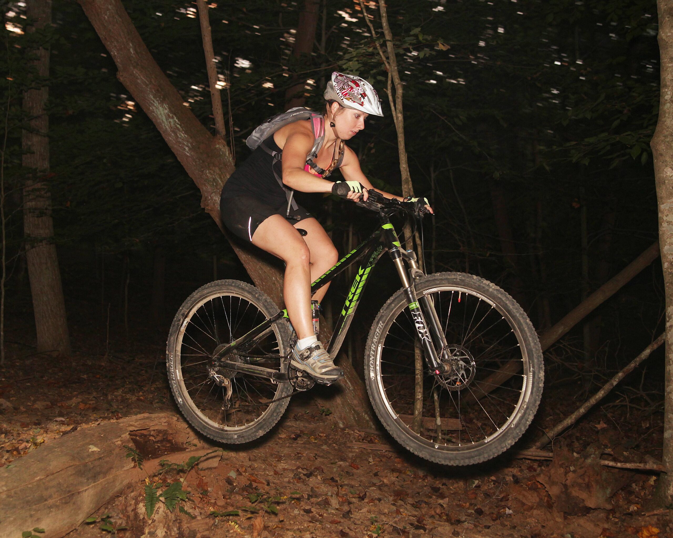 A mountain biker performing a jump over a log in a wooded area at night. The cyclist is wearing a helmet and sporting gear, with a focused expression as they navigate the terrain. Trees and underbrush are visible in the background, illuminated by the ambient light. Green River mountain bike trail.