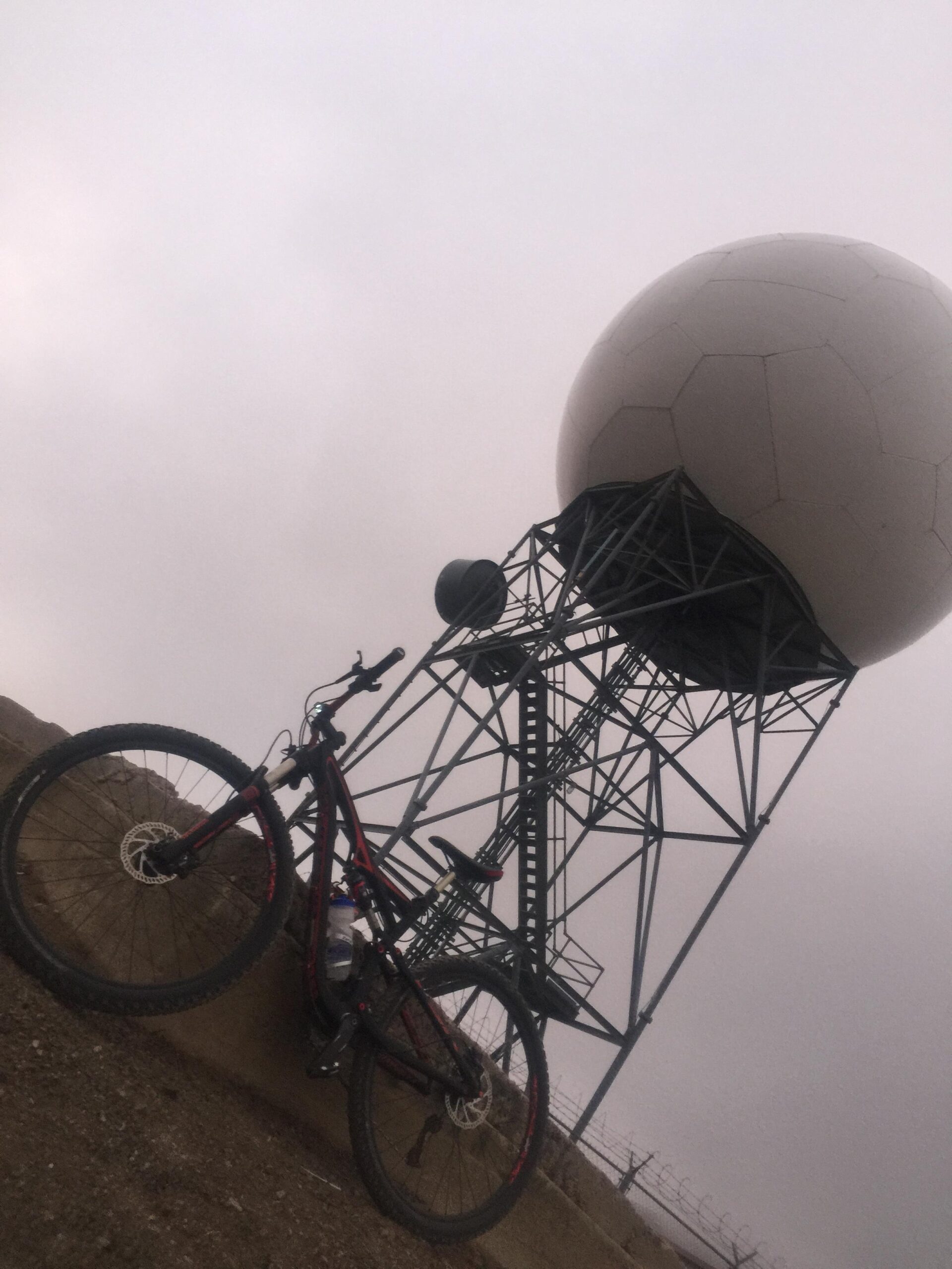 Specialized Camber 29: A mountain bike positioned beside a large, round radar dome on a metal tower, set against a cloudy sky. The bike is black with red accents and features a water bottle mounted on the frame. The terrain around the tower appears rocky and barren.