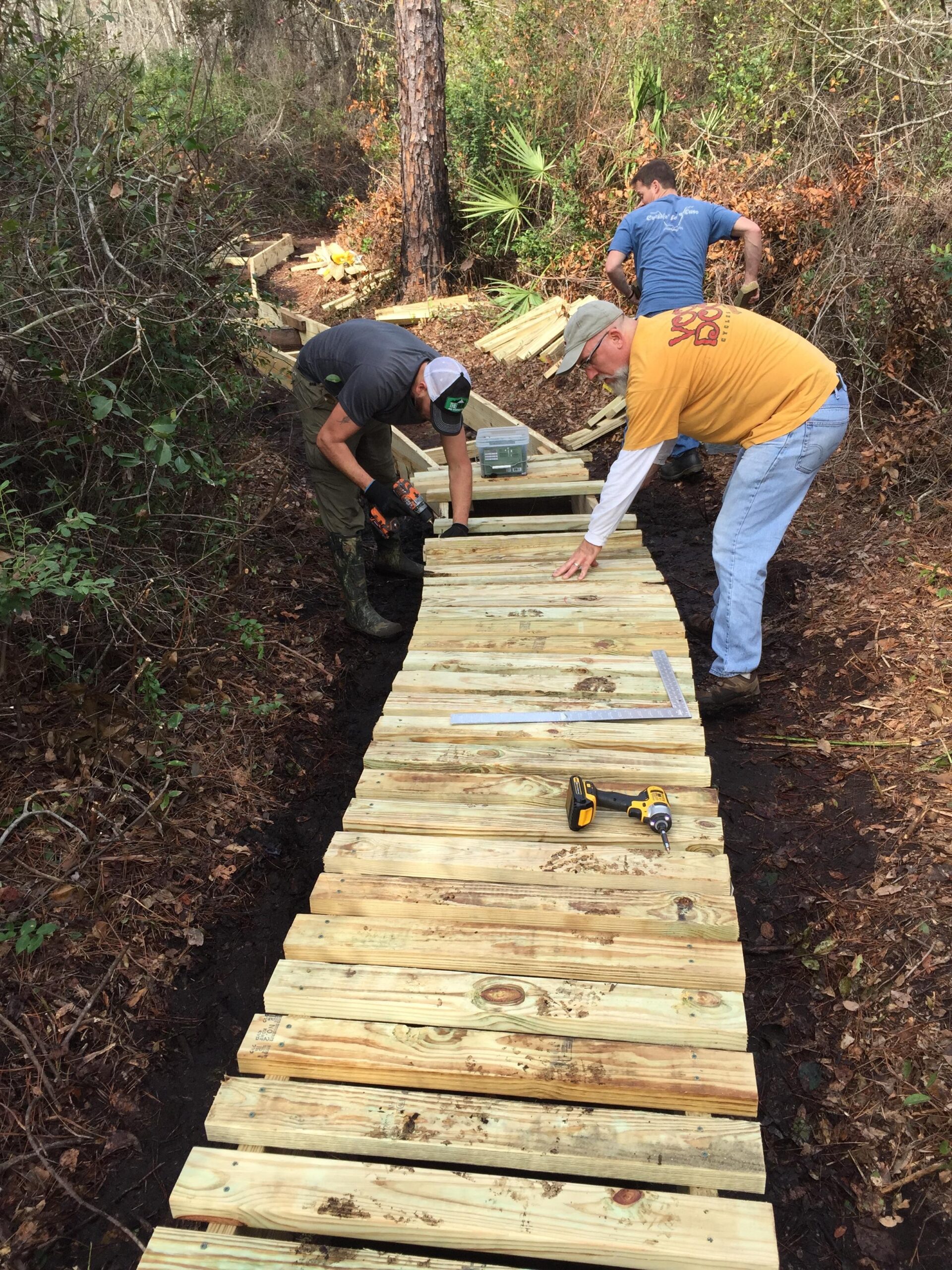 Three individuals are working together to build a wooden path in a wooded area. The scene shows them assembling planks of wood, with tools like a drill and a square visible on the ground. The pathway is framed by lush greenery and fallen leaves, suggesting an effort to enhance the accessibility of the trail. Nocatee mountain bike trail.