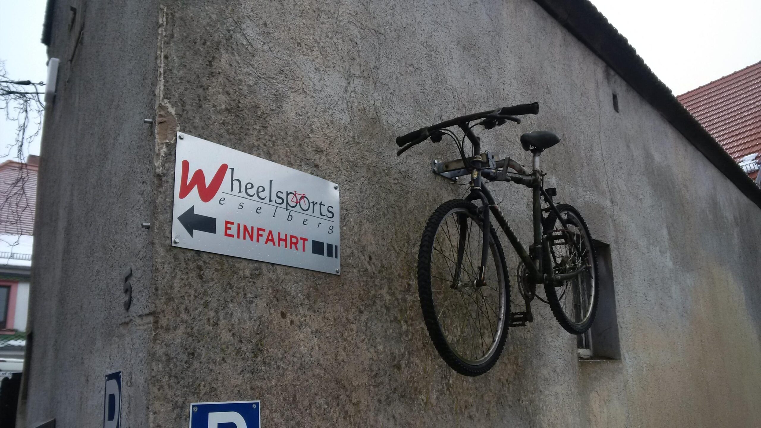 A bicycle mounted on a wall next to a sign that reads "Wheelsports Weselberg" with an arrow pointing to the left indicating the entrance. The wall is textured and has some wear, with a building and a parked car visible in the background.