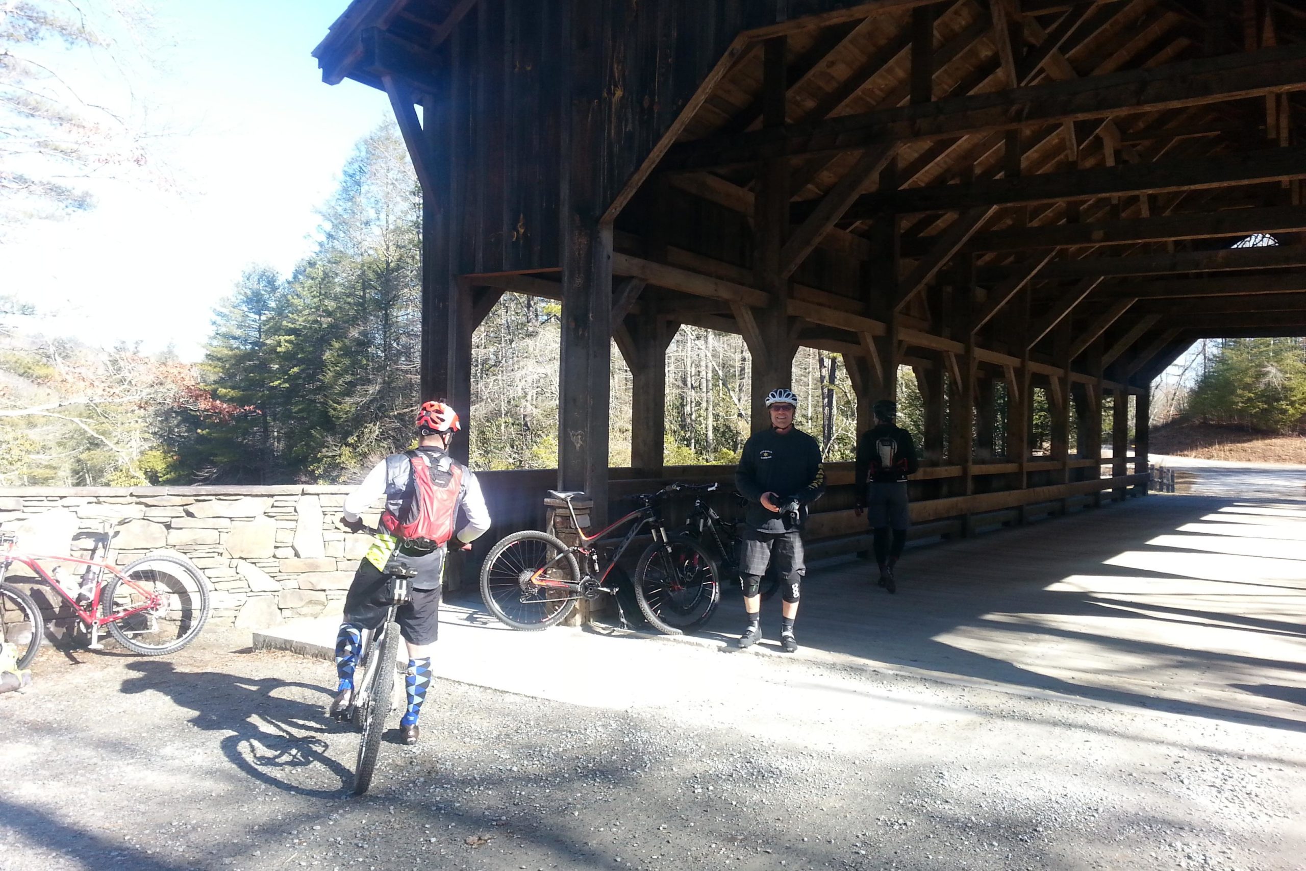 Three mountain bikers gather under a wooden shelter, surrounded by lush greenery. Two bikes are parked nearby, with one biker standing with a bike, while another biker takes pictures. Sunlight casts shadows on the ground, creating a warm atmosphere. The scene captures a moment of camaraderie and outdoor adventure. DuPont State Recreational Forest mountain bike trail.