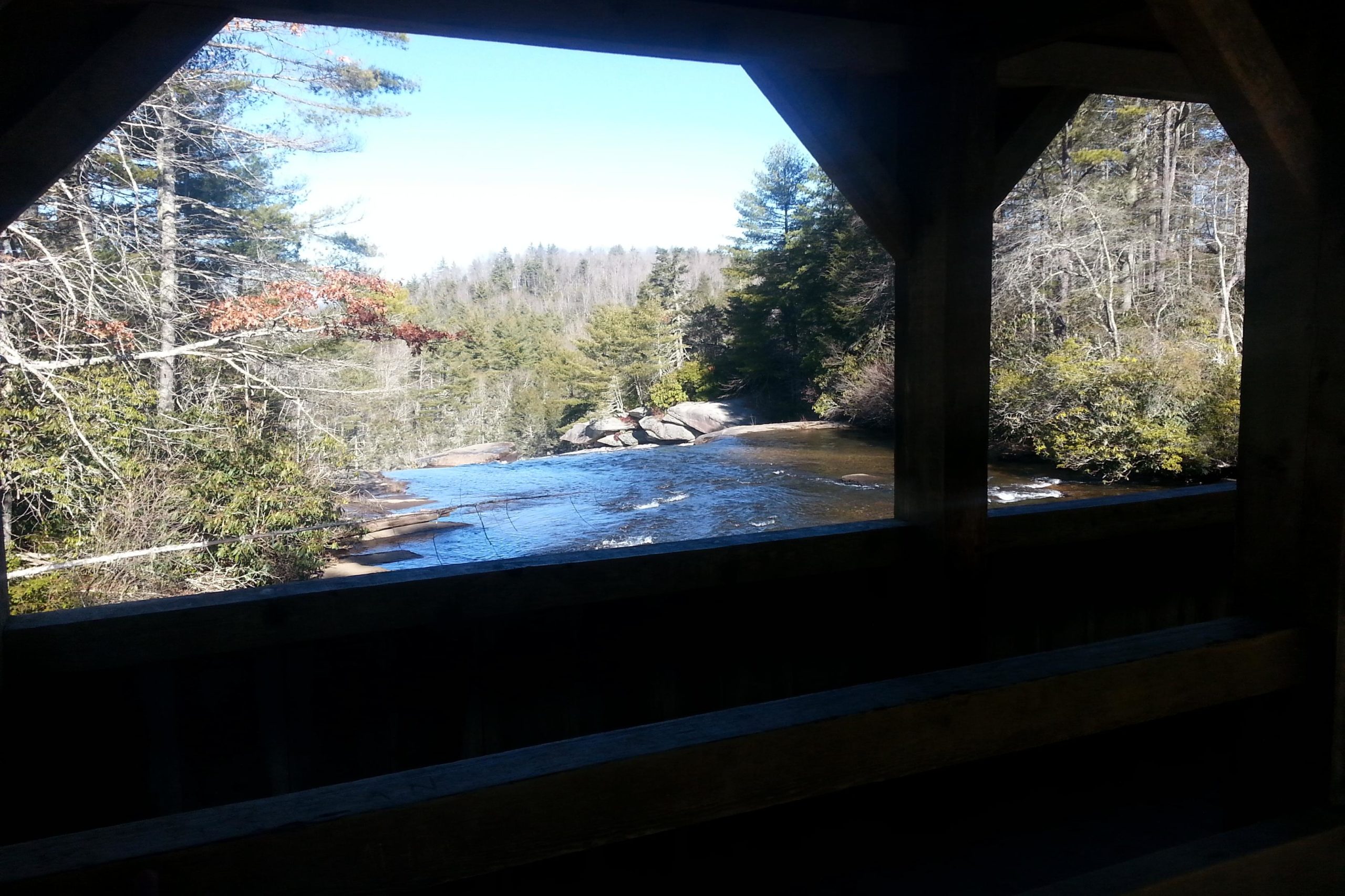 View of a tranquil river flowing between trees, framed by a wooden structure. The scene captures the natural beauty of a forested landscape with a clear blue sky above. DuPont State Forest mountain bike trail.