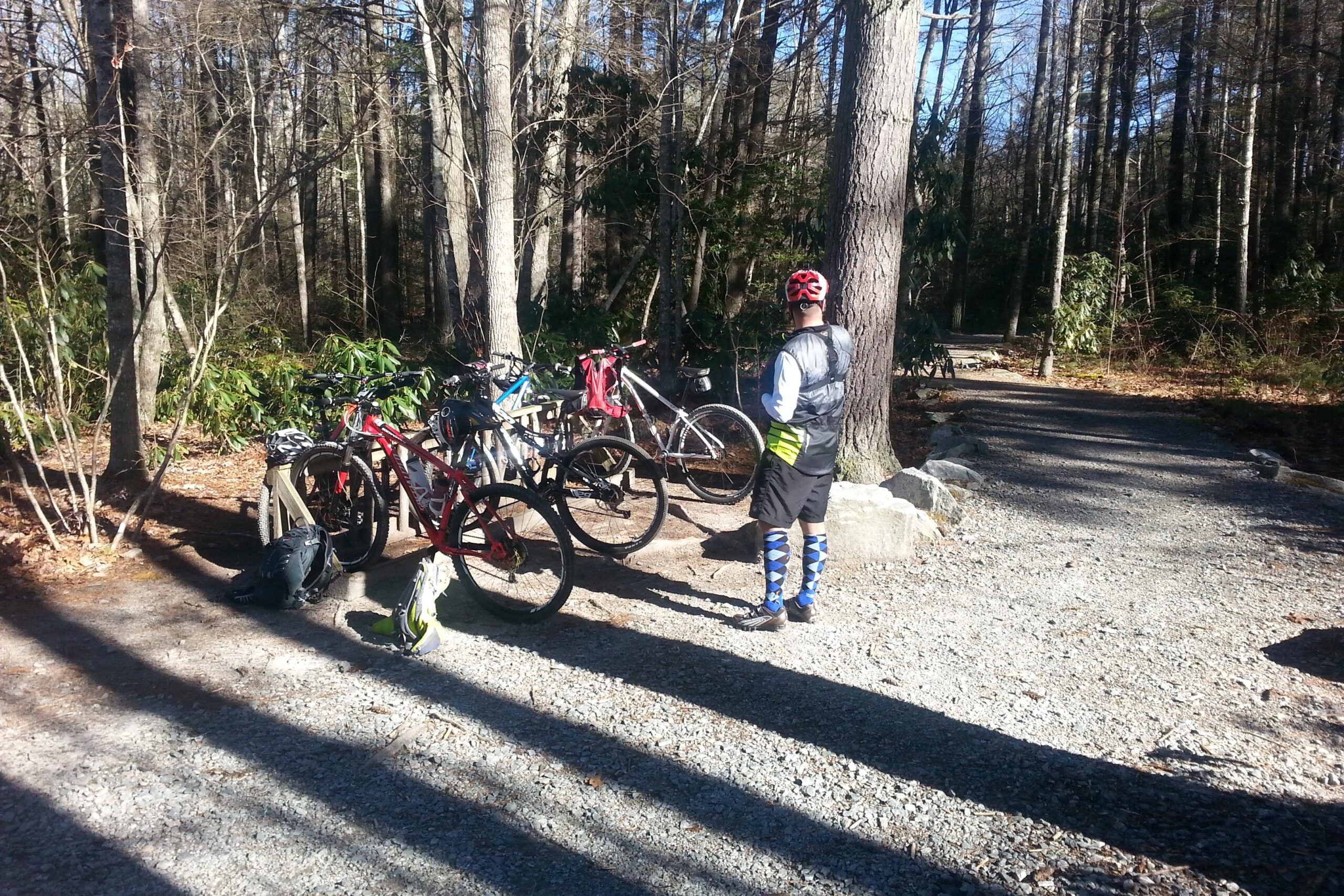 A cyclist in a black vest and colorful socks stands next to several parked mountain bikes on a gravel path surrounded by trees. Sunlight filters through the branches, casting shadows on the ground. DuPont State Forest mountain bike trail.