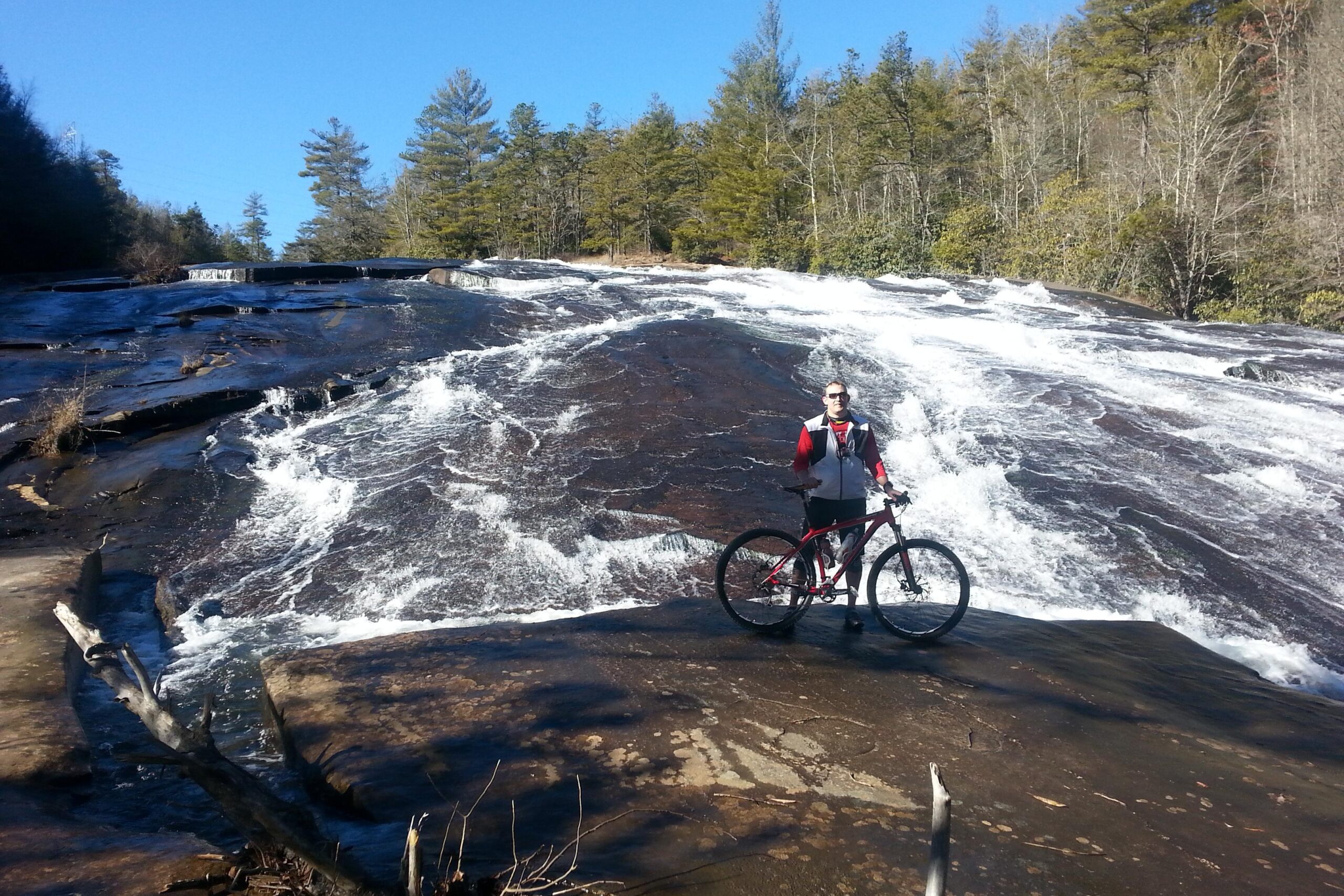 A cyclist stands next to a mountain bike on a rocky surface with a waterfall cascading in the background. The sky is clear and blue, and the surrounding area is filled with trees. Water flows over the rocks, creating a scenic outdoor environment. DuPont State Recreational Forest mountain bike trail.