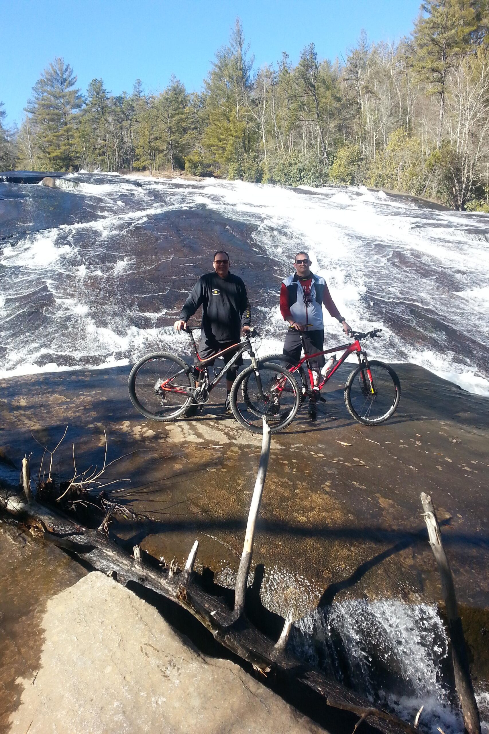 Two cyclists stand on a smooth rock surface near a flowing waterfall. They are posing with their mountain bikes, surrounded by lush trees and clear blue sky. The water cascades down the rocks in the background, creating a scenic outdoor setting. DuPont State Recreational Forest mountain bike trail.