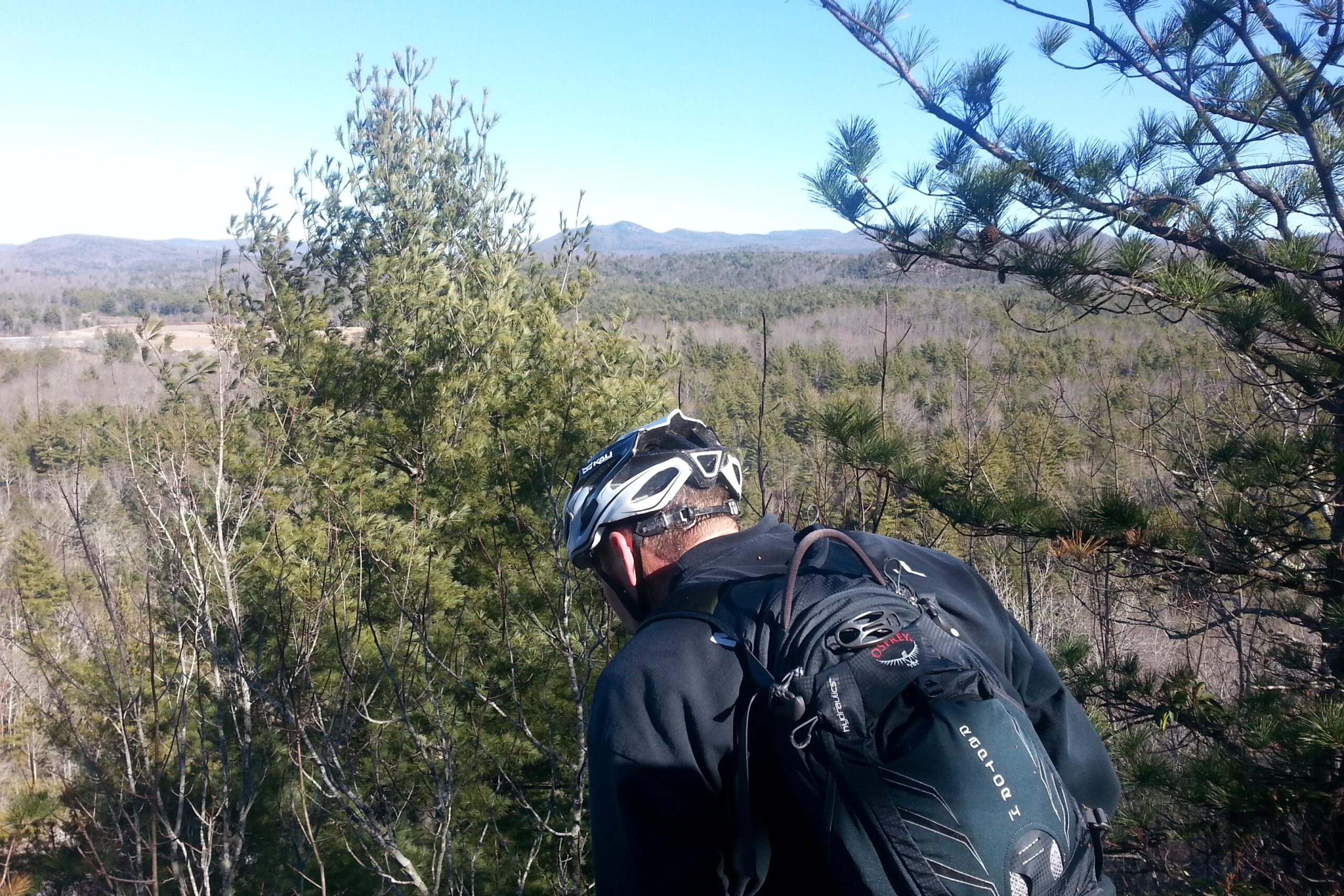 A person wearing a bike helmet and a backpack is seen from behind, overlooking a scenic view of rolling hills and trees under a clear blue sky. DuPont State Forest mountain bike trail.