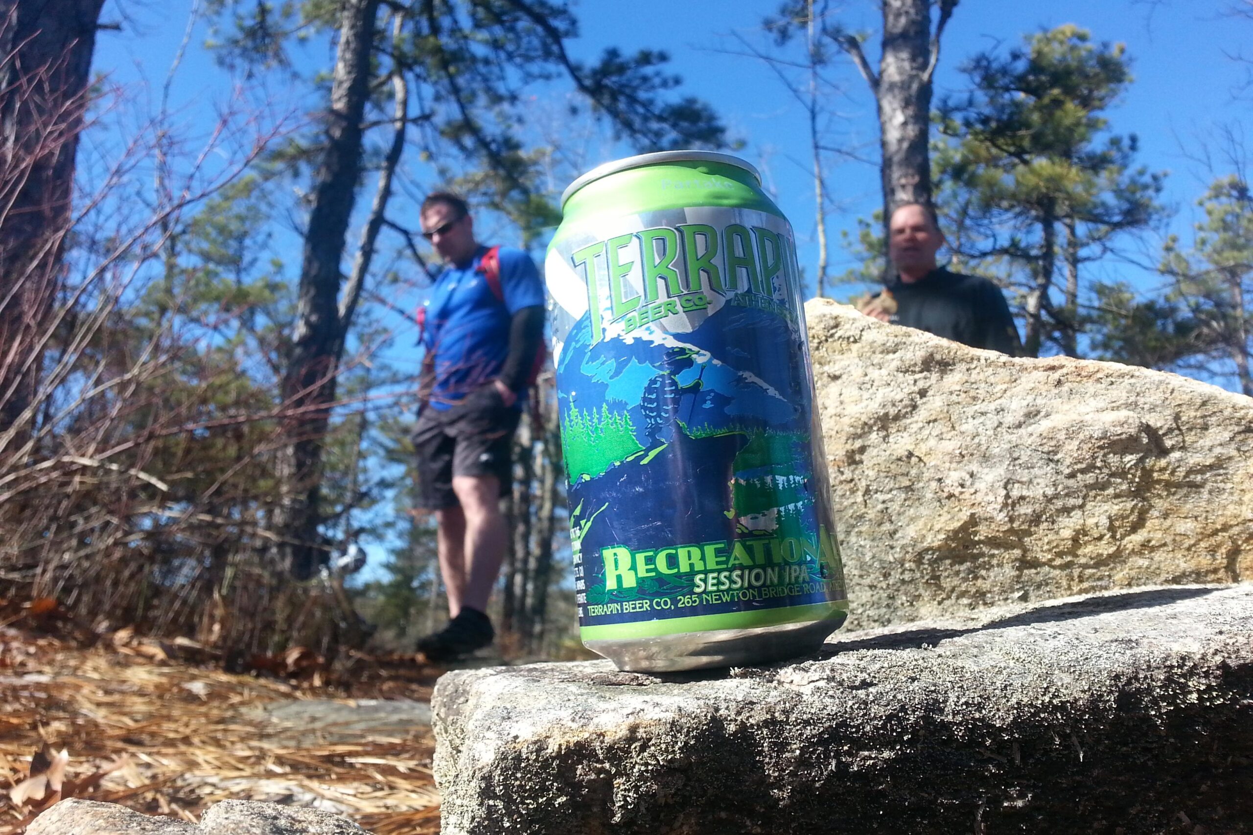A vibrant green can of Terrapin Recreation Session IPA rests on a rock in a forest setting, with tall trees and a clear blue sky in the background. Two hikers, dressed in outdoor gear, are visible in the distance, engaged in their surroundings. The scene suggests a day of outdoor adventure and enjoyment. DuPont State Recreational Forest mountain bike trail.