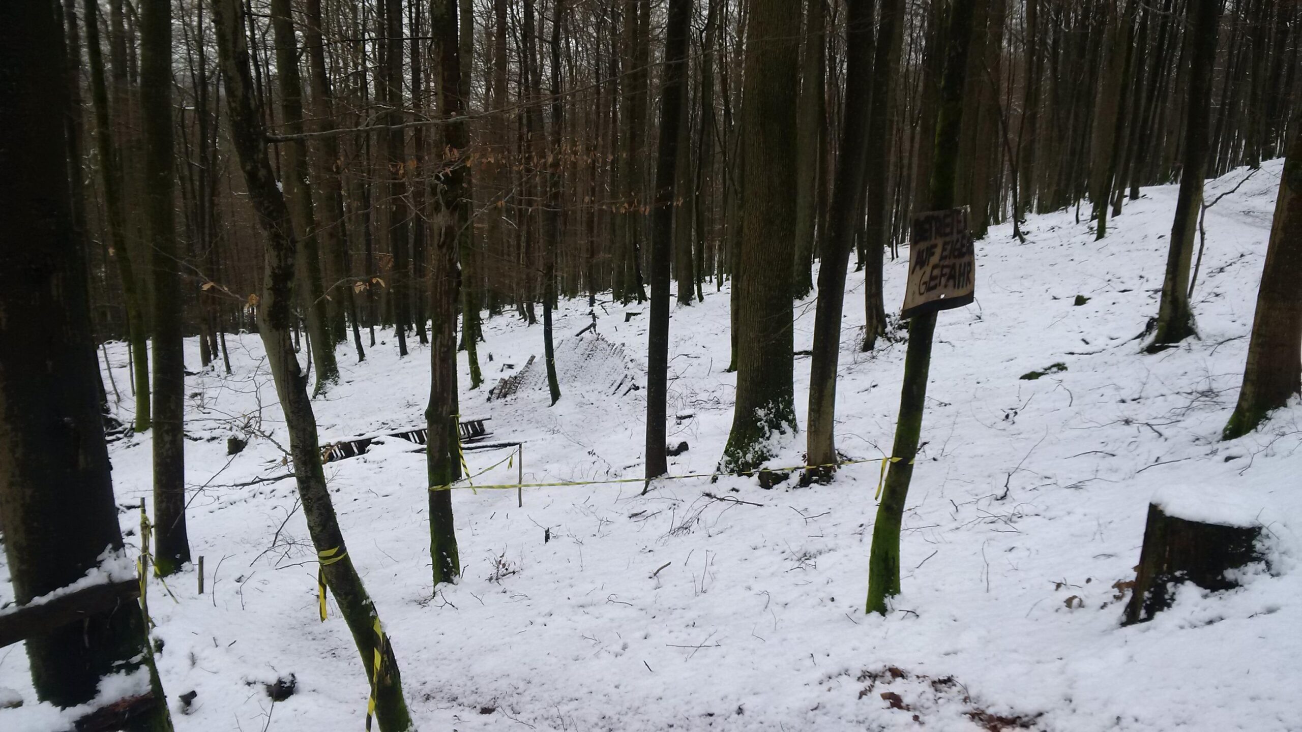 A snowy forest scene with tall, bare trees. The ground is covered in a thick layer of snow, and a wooden sign is partially visible, warning of danger. Yellow tape can be seen marking off a path or area in the forest. Technikparcours mountain bike trail.