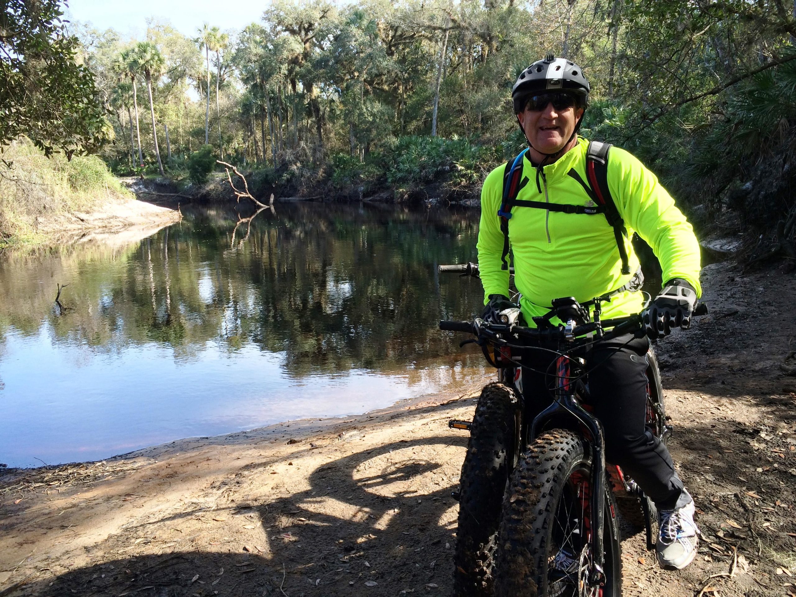 A cyclist wearing a bright yellow long-sleeve shirt, gloves, and a helmet stands beside a mountain bike with wide tires near a calm river. The scene includes lush greenery and palm trees in the background, reflecting in the water. It's a sunny day in a natural outdoor setting. Little Big Econ State Forest mountain bike trail.
