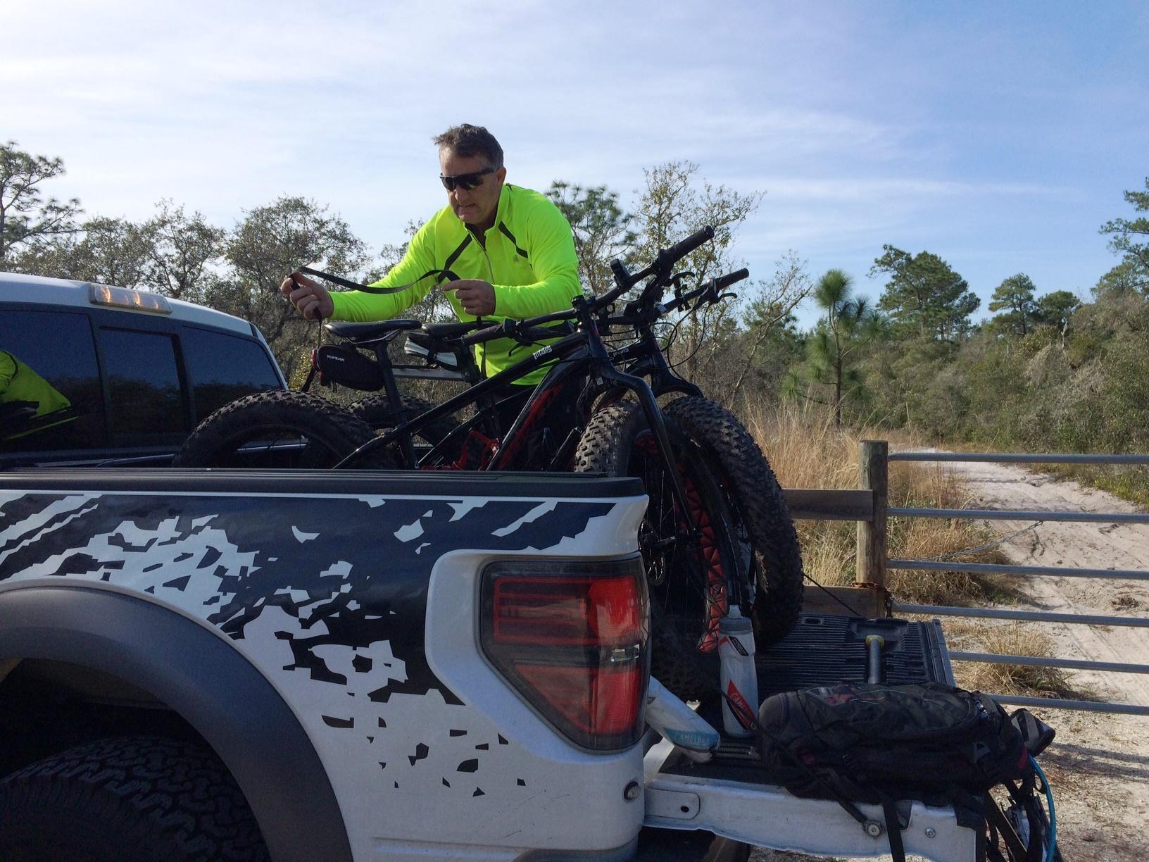 A man in a bright yellow long-sleeve shirt and sunglasses secures mountain bikes in the back of a pickup truck. The background features a natural area with trees and a sandy pathway. The truck has a patterned design on its bed, and various biking gear is visible. Little Big Econ State Forest mountain bike trail.