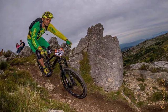Mondraker Foxy XR: A mountain biker navigating a rocky trail, wearing a green and yellow cycling jersey and helmet. The biker is leaning into a turn, with large boulders and a scenic landscape visible in the background, under a cloudy sky.